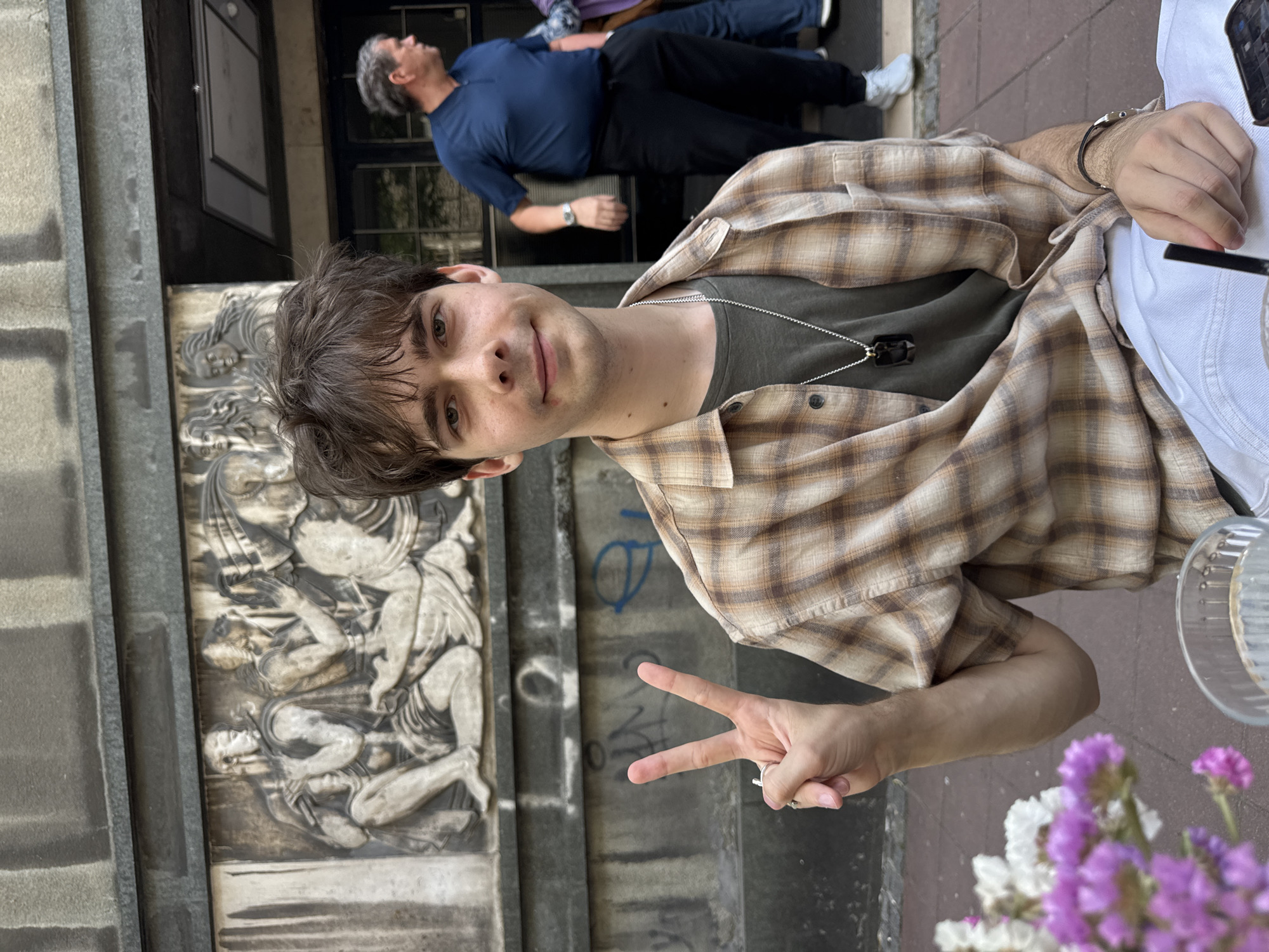 A young man with brown hair stands in front of a building featuring a detailed stone carving, possibly in Belgrade, Serbia. He wears a green t-shirt under a tan and brown plaid shirt, a necklace, and a ring on his left hand, holding up a peace sign. Another man in a blue shirt and black pants is visible further back, seemingly lying down. Flowers are in the foreground near a clear glass. The building has a dark door and gray exterior.
