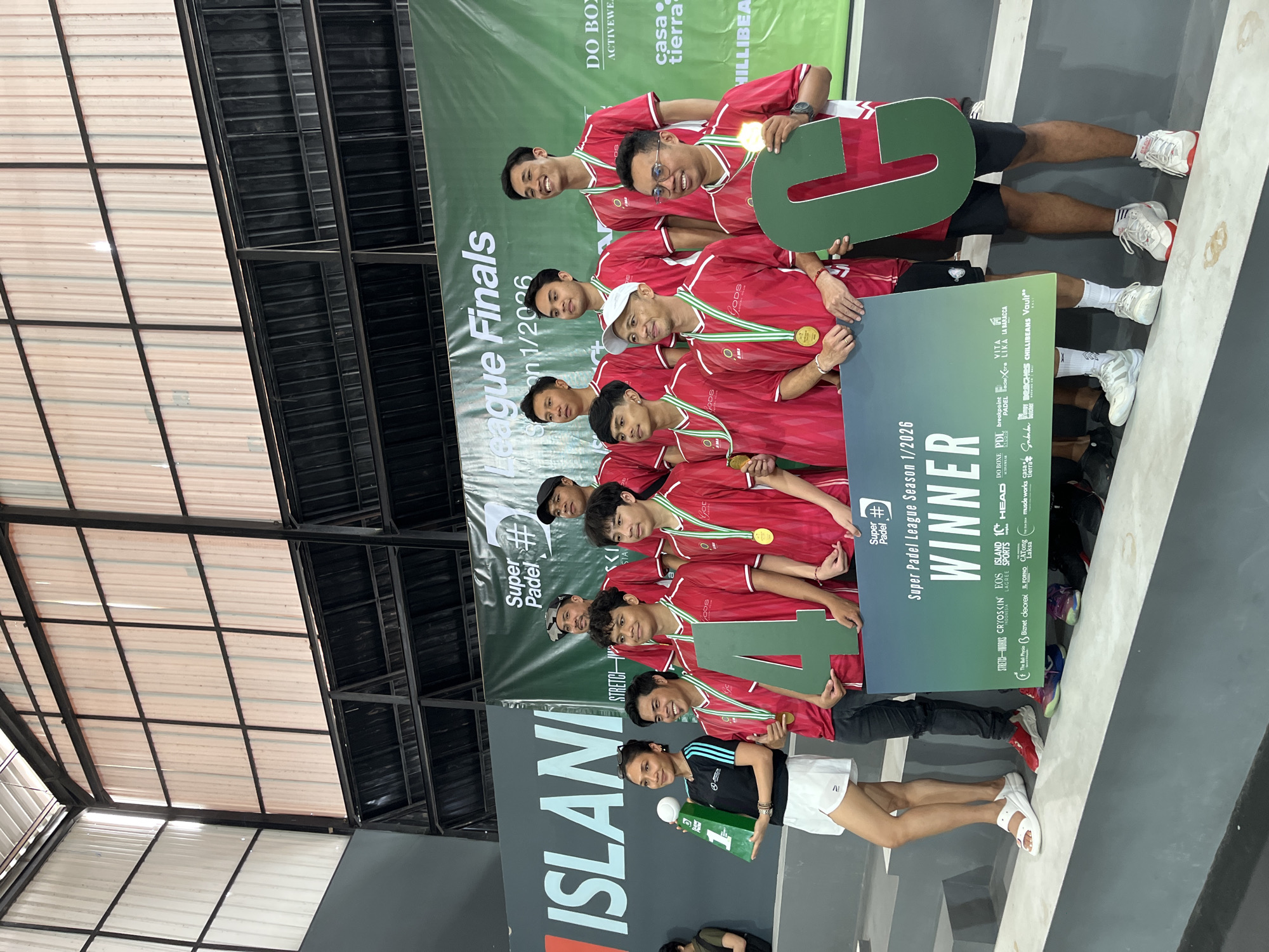 A sports team is celebrating their victory at a padel tournament held in North Kuta, Indonesia. There are 10 male athletes wearing red jerseys and gold medals around their necks. They are holding up a banner that says "Super Padel League Finals Season 1/2026 WINNER". Two of the athletes are holding the digits "4" and "6," implying a '46' for their team. An athlete in a black shirt and white shorts is holding a trophy shaped like the number "1". The backdrop includes sponsors such as "DO BOX ACTIVEWEAR" and "MILLIBEA", along with other brand names. The event appears to be taking place indoors with a visible corrugated metal roof structure.