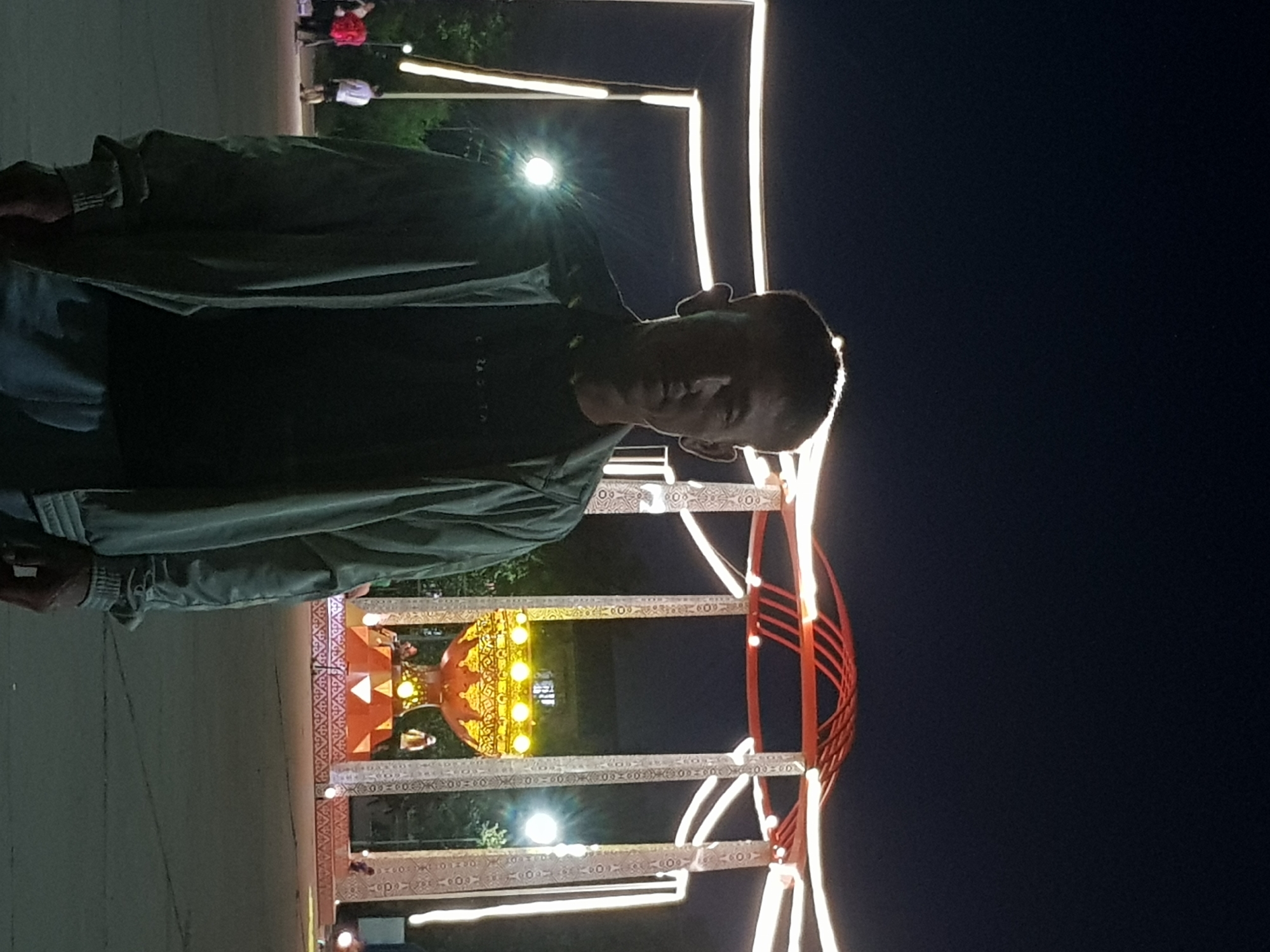 A medium shot shows a young man standing in front of a large, illuminated decorative structure at night. The man is facing the camera, his head slightly bowed, with a serious expression. He is wearing a dark t-shirt and a light-colored, possibly grey or blue, bomber jacket. The background features the ornate structure, which appears to be a pavilion or monument, with intricate geometric patterns and bright yellow lights. Several white lights outline the structure and its supports, creating a luminous effect against the dark night sky. In the distance, other figures can be seen, some illuminated by the lights, suggesting a public space or event. The ground is paved. The presence of the illuminated structure suggests it might be a landmark or part of a festival. This scene is identified as being in the city of Kurchatov, Kazakhstan.