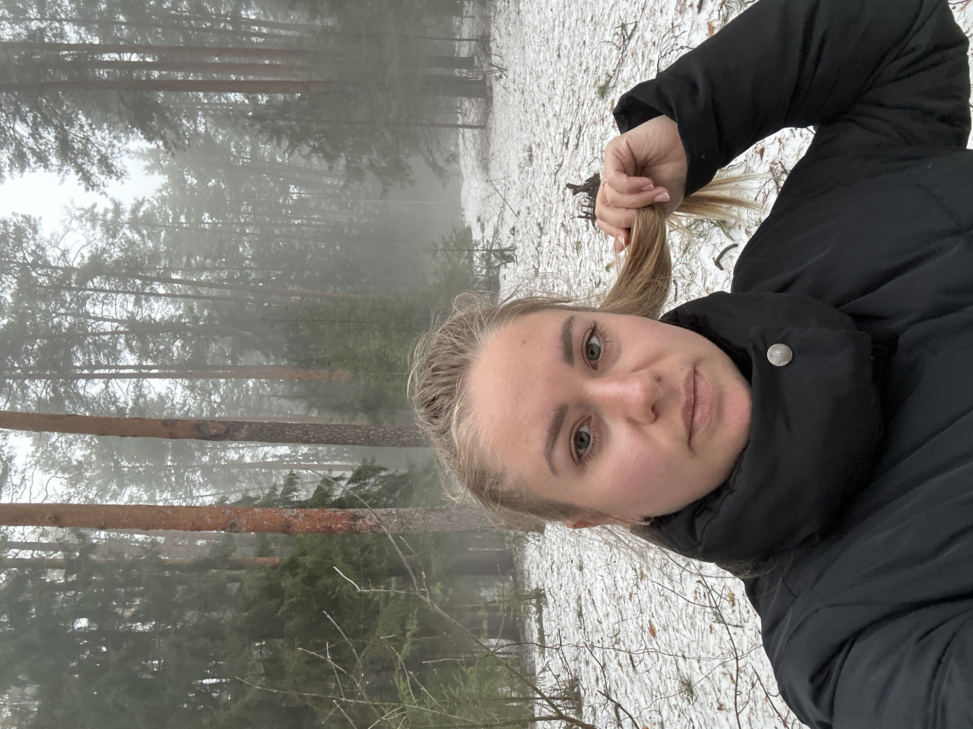 A young woman is taking a selfie in a snowy, foggy forest. She is wearing a black puffer jacket. She is holding a strand of her light brown hair. Tall pine trees surround her, and a light dusting of snow covers the ground. The location appears to be a wooded area, possibly a park, near or within the city of Chaikovsky, Russia.