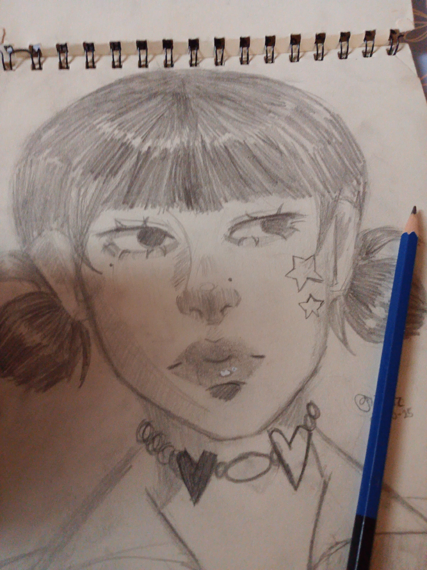 A graphite sketch of a young person's face is depicted in a sketchbook. The sketch is close-up, focusing on the head and shoulders. The subject has bangs, pigtails, and is wearing a necklace with heart and circular charms. There are also two stars drawn on the right cheek. A blue pencil is positioned vertically along the right side of the image, partially obscuring the sketch. The background is the surface of the sketchbook, with spiral binding visible at the top. The sketch is rendered with soft shading and linework. There is a date scribbled in the lower right corner of the sketchbook page. The location in the city of Kiblawan, Philippines, is not discernible from the provided image.