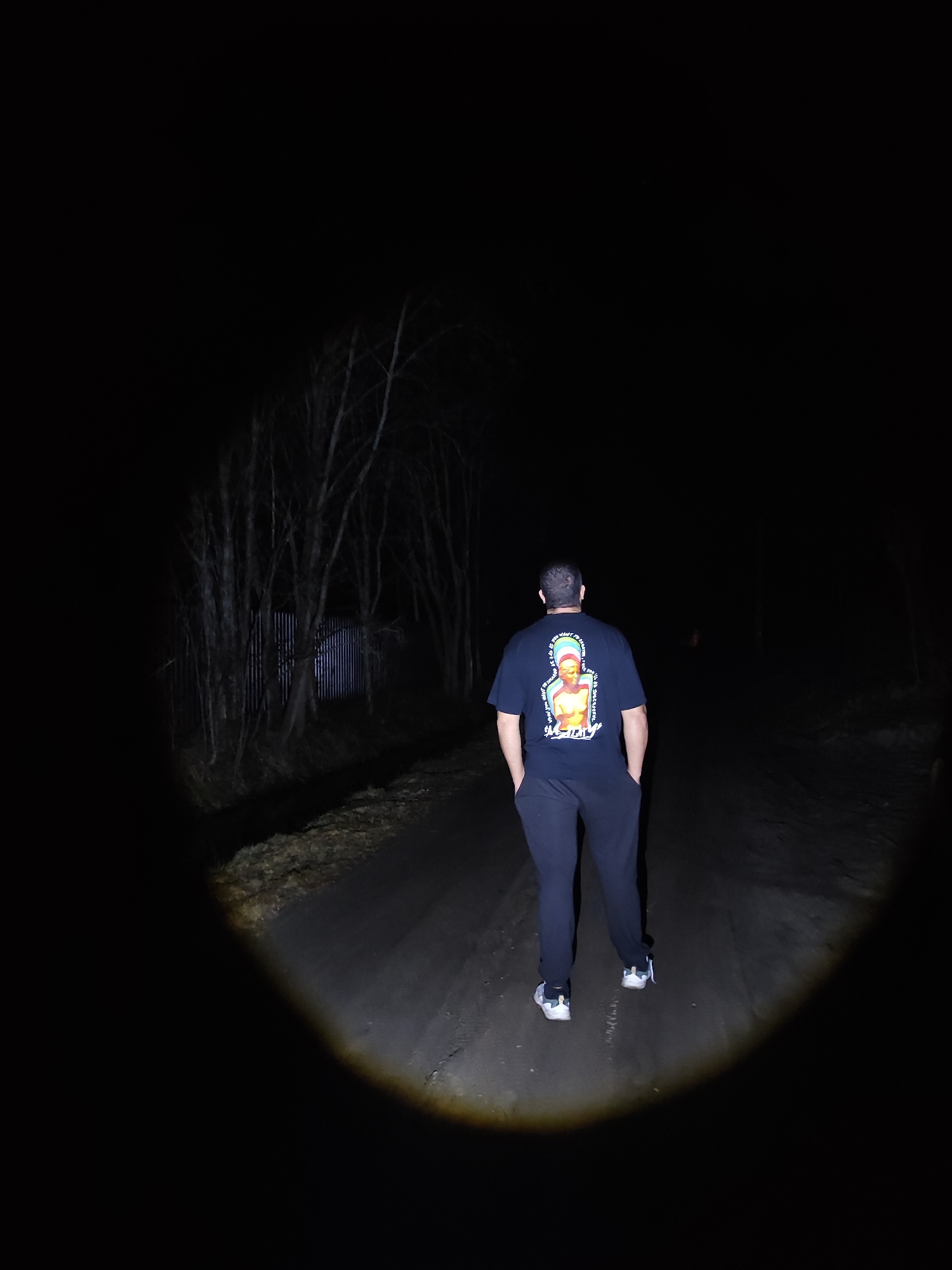 A person stands on a road in a dark, wooded area at night. The person is wearing a black t-shirt with a design on the back, black pants, and white shoes. The person has their hands in their pockets. A beam of light shines from the lower center of the image, illuminating the road and the person. The sides of the road are lined with trees and a corrugated metal fence. In the distance, a faint light is seen. The city and location are unknown.