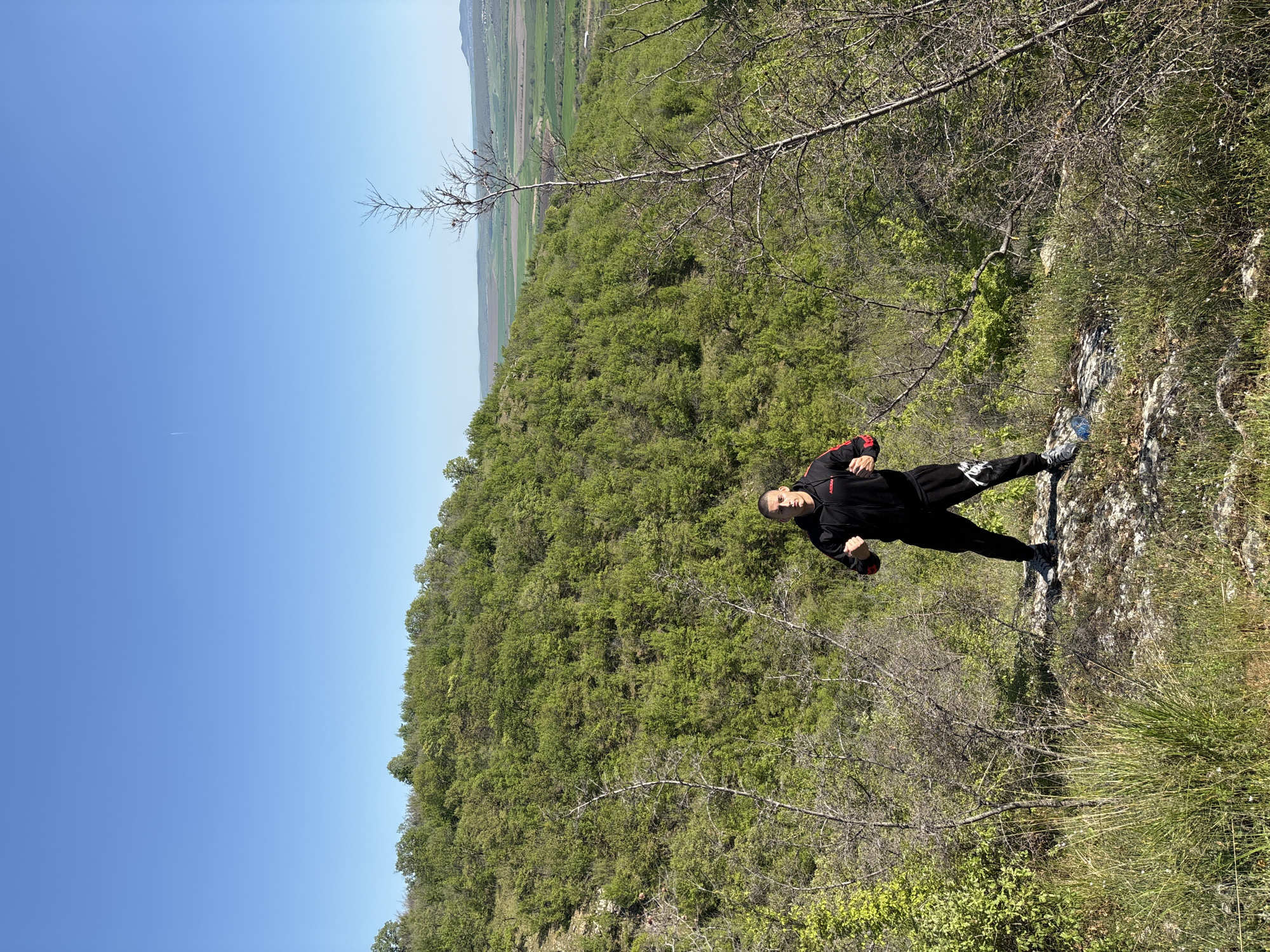 The image shows a man standing on a rocky outcrop. He is wearing a black tracksuit with red detailing. He is posing with one arm raised. The background features a dense green forest and a blue sky. In the distance, flat agricultural land is visible. The image was taken in the Goritsa region of Bulgaria, suggested by the foliage and terrain.