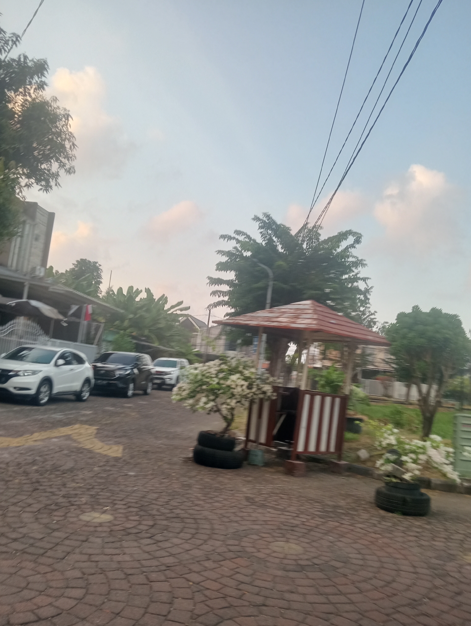 The image shows a paved driveway with several cars parked on it. There is a small gazebo with a red roof and white striped sides. In the foreground, there is a tree with white flowers planted in what appear to be old tires. In the background, there are houses and trees. The sky is light blue with some clouds. The image appears to have been taken in Surabaya, Indonesia, given the style of the houses and the vegetation.