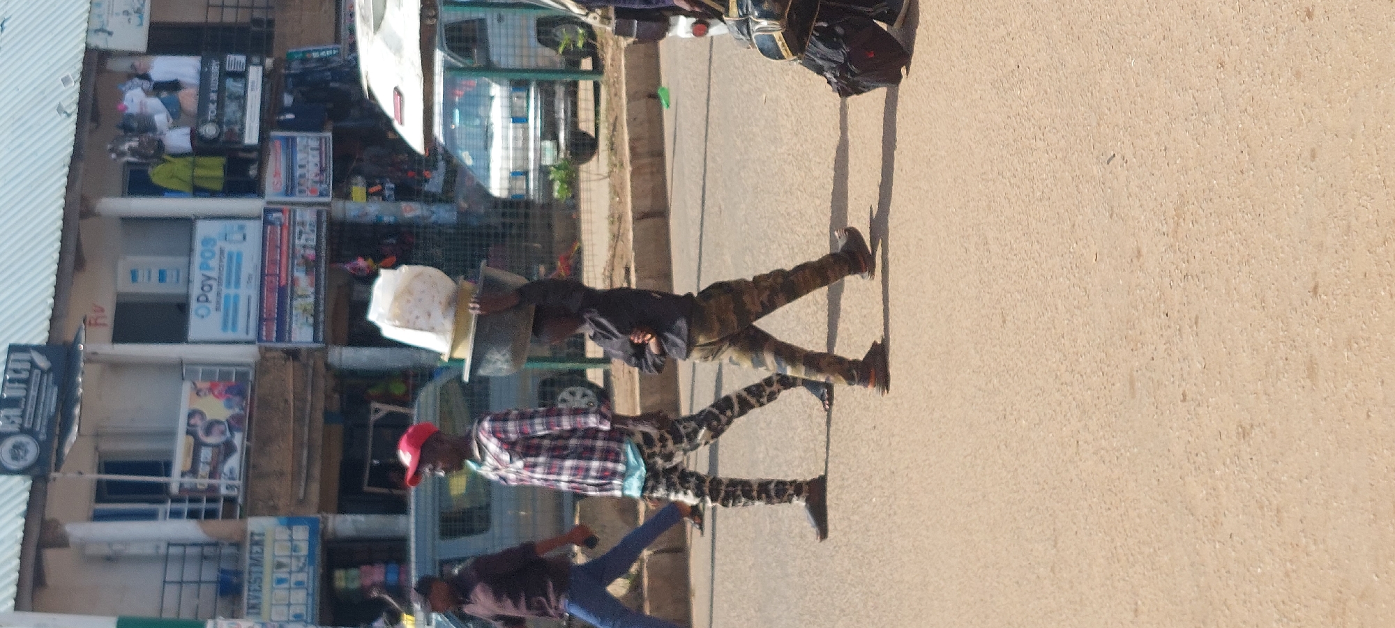 A young boy is walking across a paved road, balancing a stack of items on his head, which include two bowls and a white bag. He is wearing a dark long-sleeved shirt and camouflage pants. To his left, a man in a red cap and a plaid shirt walks ahead, wearing patterned pants. To the far left, another man in a dark shirt and blue jeans walks in the same direction. In the background, there is a row of buildings with various shop signs, including "INVESTMENT," "O Pay POS," and "TOK IN LUXURY." A light-colored van is parked behind the boy, and another vehicle is partially visible to the far right. The ground is a light brown asphalt, and there is a curb running along the side of the road.