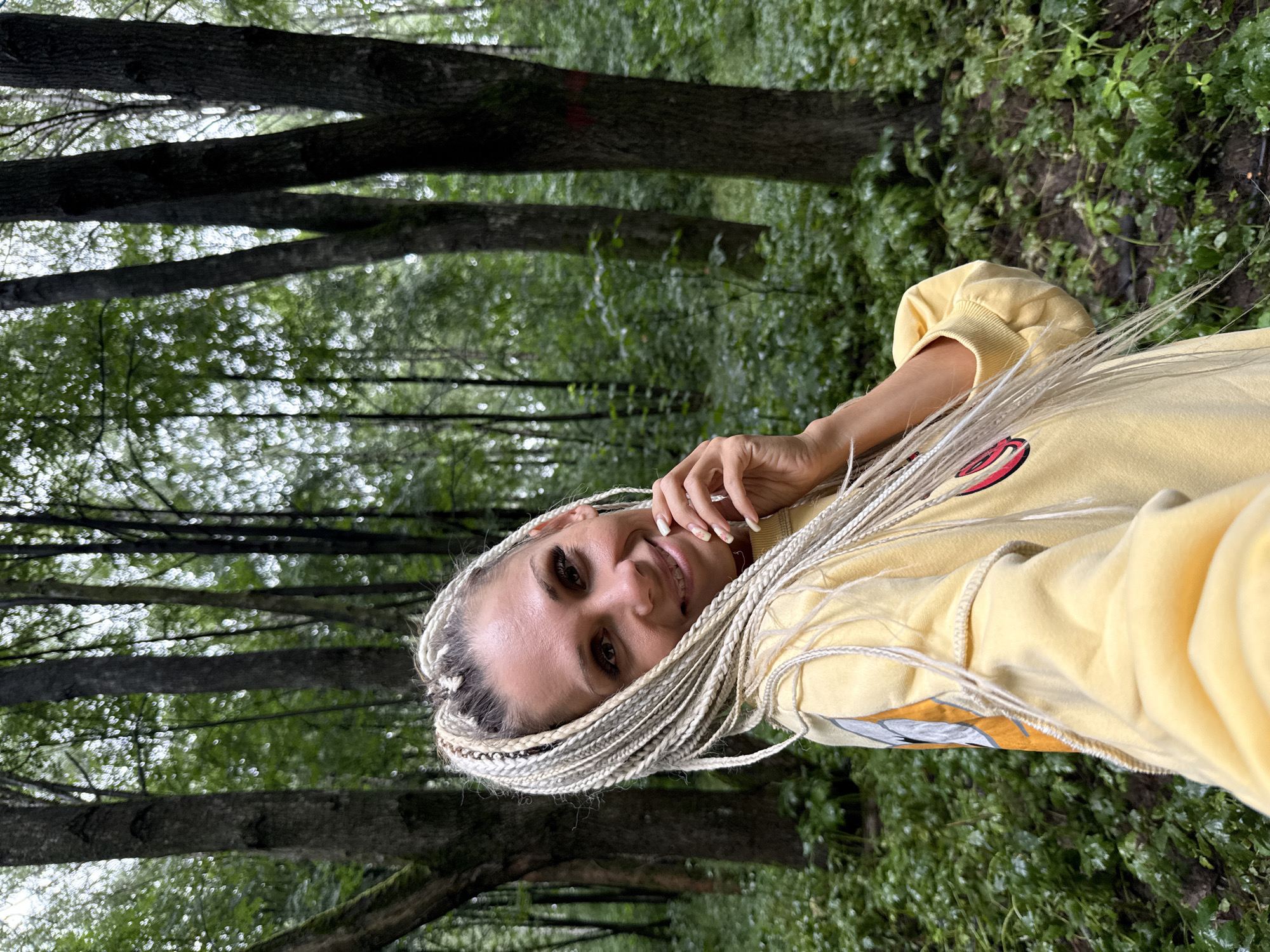A woman with blonde braided hair smiles at the camera in a forest. She is wearing a yellow sweatshirt. The background is a dense forest with tall trees and green foliage. The lighting suggests it is daytime. The location is specified as the city of Dumino, Russia.