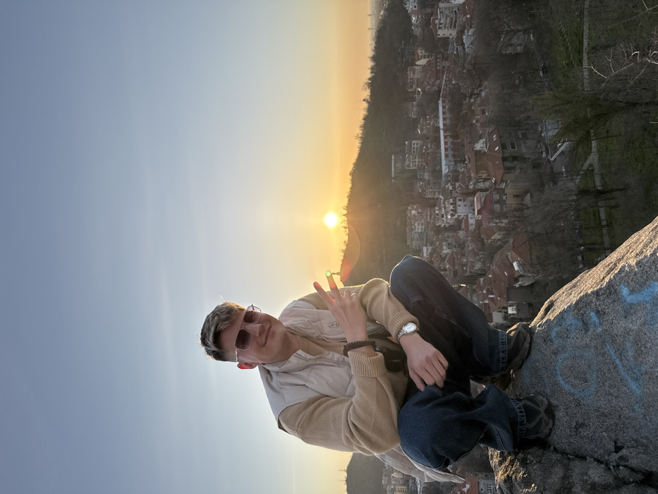 A male individual is depicted squatting on a large grey rock on an elevated vantage point overlooking the city of Plovdiv, Bulgaria. The subject, with light-colored hair and wearing sunglasses, a beige long-sleeved top, a light-colored puffer vest, dark blue jeans, and dark athletic shoes, is smiling directly at the camera. A silver-toned watch is present on his left wrist, and a dark beaded bracelet is on his right wrist. His right hand is raised, forming a peace sign. The rock he is positioned on features faint light-colored markings.
The background presents a panoramic view of Plovdiv, characterized by numerous buildings with red-tiled roofs and multi-story structures, consistent with residential and urban areas. Sparse, leafless trees and some green foliage are interspersed throughout the cityscape. The sun, visible as a bright orange orb, is setting behind a distant, tree-covered hill, casting an orange-yellow glow across the horizon. The sky transitions from a clear pale blue overhead to warmer hues near the sun. The scene is situated on one of Plovdiv's elevated hills, providing an unobstructed view of the city below and surrounding topography.