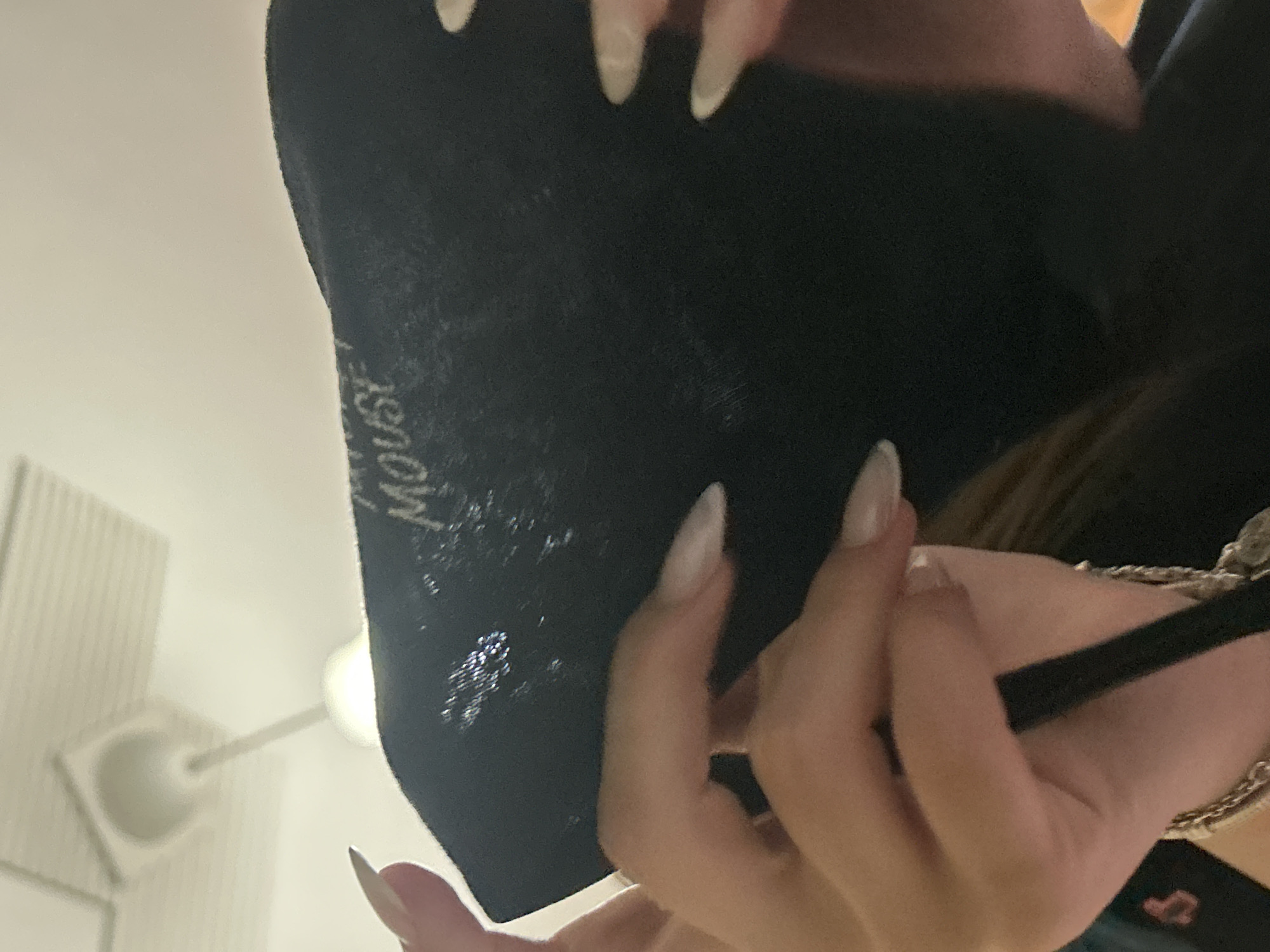 An individual is depicted indoors, holding a black fabric item to obscure their face. The fabric bears white text, partially visible as "MOUSE," suggesting the word "MICKEY MOUSE." A glossy or glittered texture is also present on the fabric. The individual's hands are visible, adorned with long, light-colored, almond-shaped fingernails. A silver-toned chain bracelet is visible on one wrist. A black strap or cord is held in the left hand. In the background, a white ceiling with a rectangular grid pattern is visible, from which a white, inverted dome-shaped light fixture hangs via a long white rod. The specific location in the city of Plovdiv, Bulgaria, cannot be determined from the visual information presented in the image.