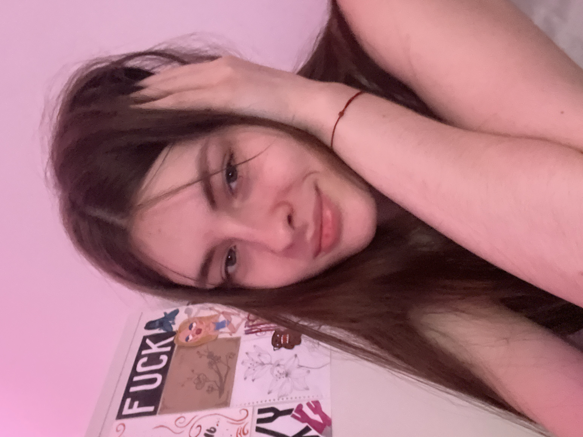 A young woman with long, dark brown hair is lying on her side, facing the camera. She has fair skin, green eyes, and a slight smile. Her right hand is resting on her head. She is wearing a thin red bracelet on her left wrist. The background is a light pink wall. There is a white object with a collage of images and the word "FUCK" on it to her right. The location cannot be determined from the image.