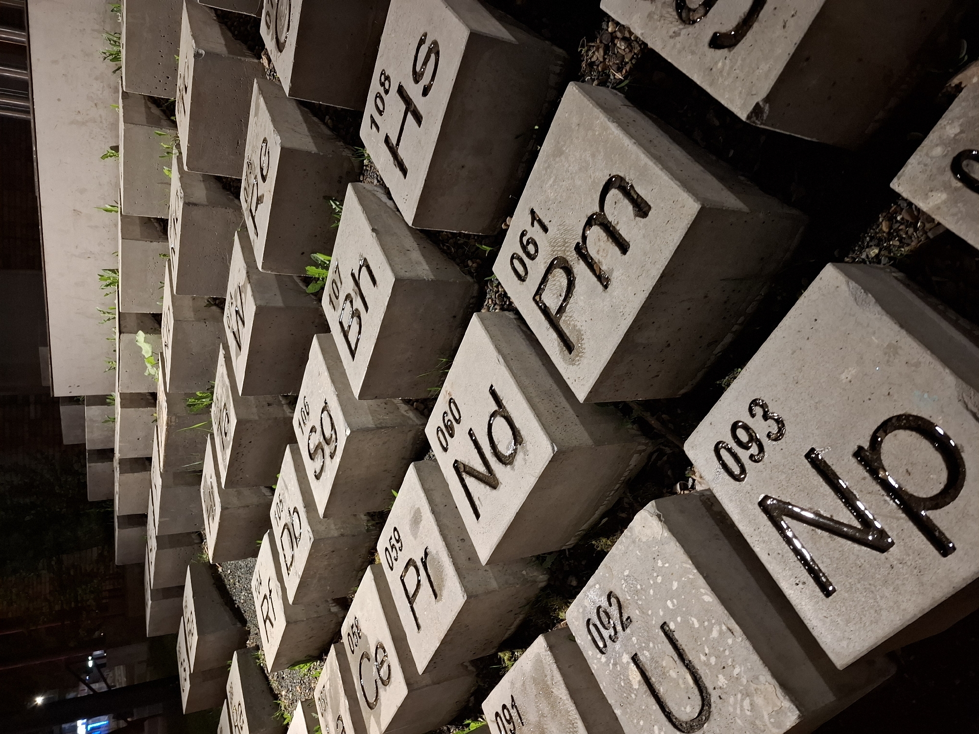 The image captures a display of concrete blocks arranged in rows, each block marked with a number and chemical symbol, reminiscent of the periodic table of elements. The blocks are weathered and positioned outdoors, as evidenced by small plants growing between them. The scene is likely an art installation or memorial in a public space. Based on the markings on the blocks, this is the "Alley of Chemical Elements," located in Glazov, Russia. The lighting suggests the photo was taken at dusk or night, with artificial lights providing illumination. The blocks are slightly angled, and the composition of the photo draws the eye along the rows of elements.