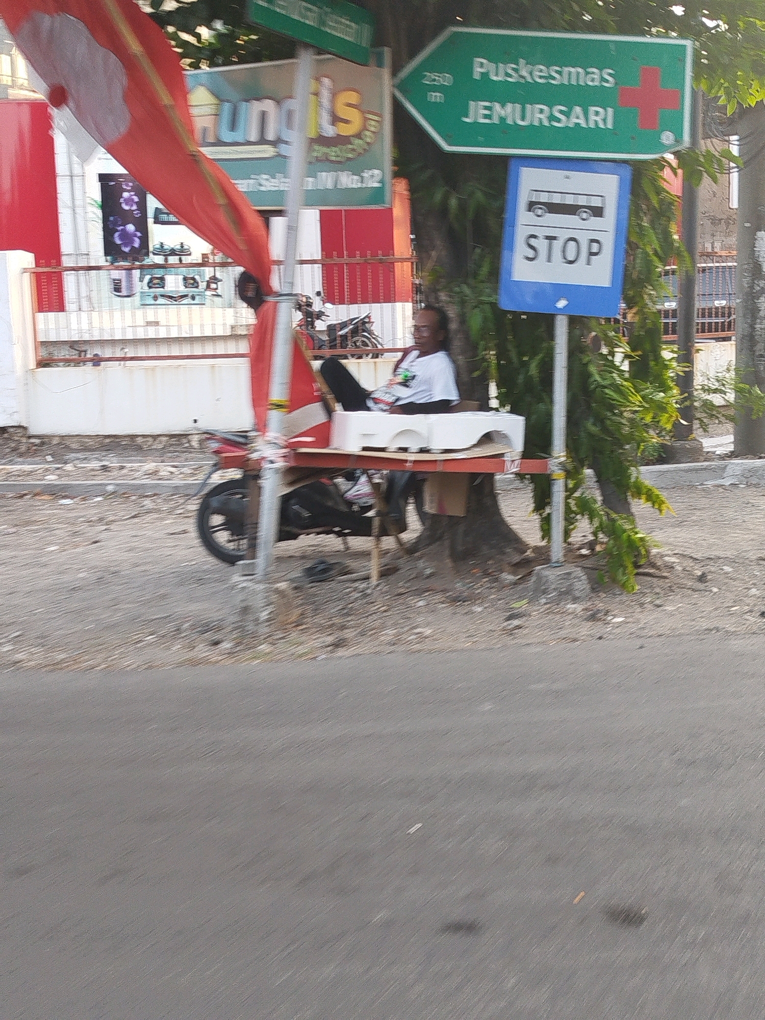 A person sits on a cart near a bus stop sign on a street in Surabaya, Indonesia. The bus stop sign indicates "Puskesmas Jemursari" and has a red cross symbol, with "250 m" written above it. Below the Puskesmas sign is a blue sign with a bus icon and the word "STOP". A red and white Indonesian flag is draped over a pole. In the background, there are buildings and trees. A motorcycle is parked on the side of the road. The ground is covered in gravel and debris.
