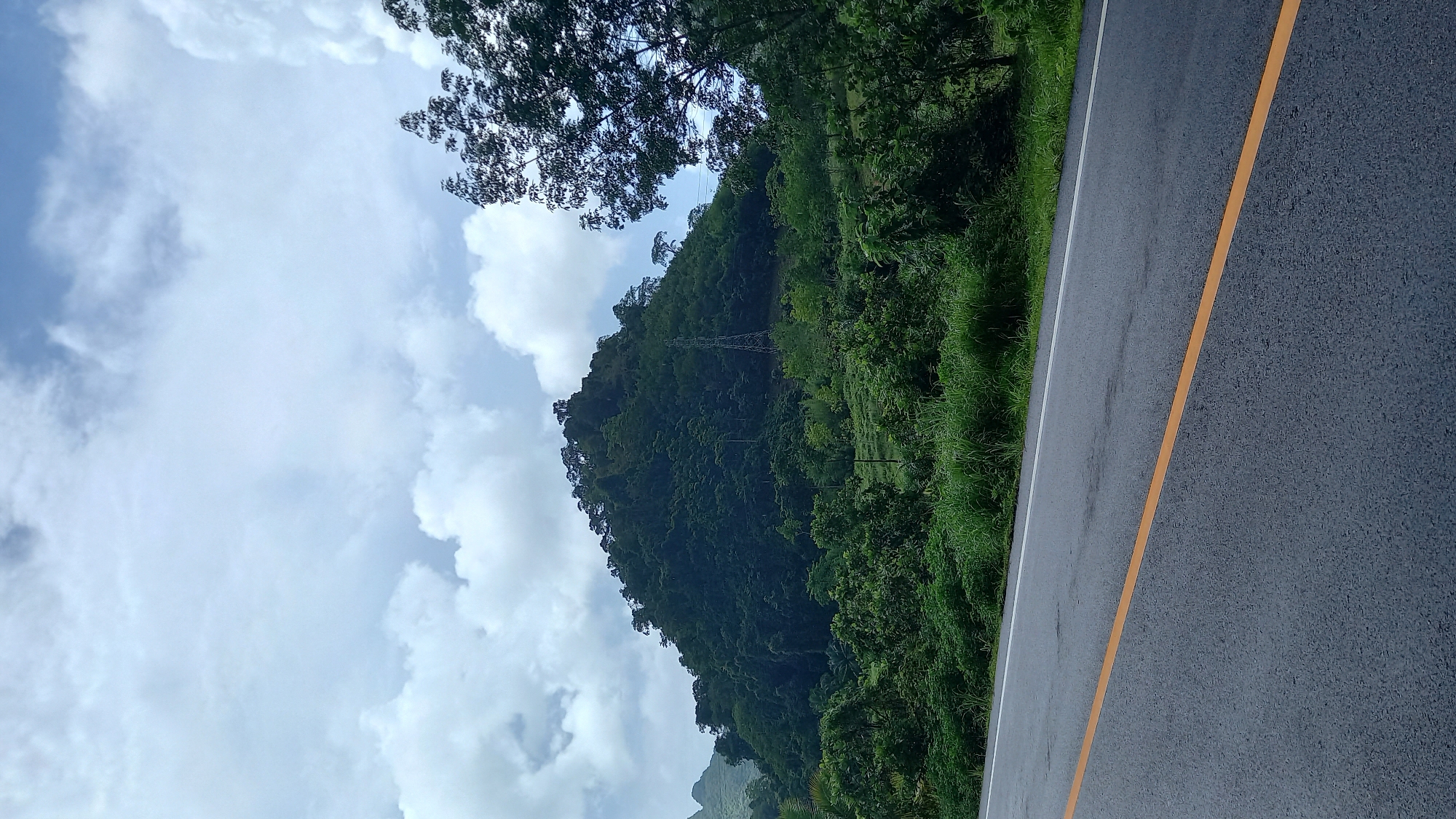 A partially obscured, green, densely vegetated mountain slope rises from the edge of an asphalt road, under a sky filled with puffy white clouds and patches of blue sky in Ban Pak Ra, Thailand. A large tree stands partially visible on the top left edge of the image. An electrical power line tower is barely visible on the mountain slope. The road has a yellow line. Grass grows on the shoulder of the road.