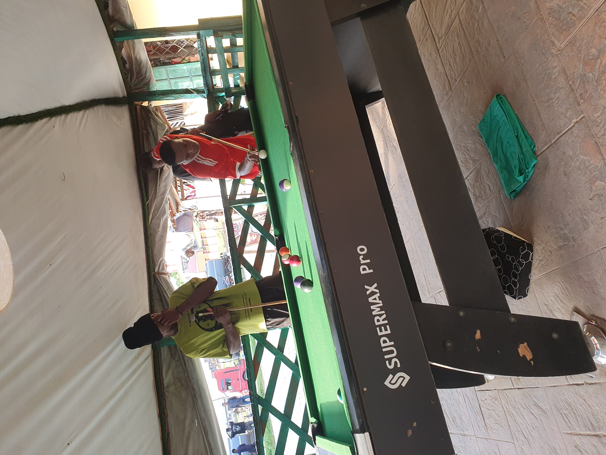Two young men are playing billiards at an outdoor venue in Lokoja, Nigeria. The man on the left, wearing a green t-shirt and a black beanie, is holding a billiard cue and appears to be observing the game. The man on the right, in a red t-shirt with white stripes, is actively playing, positioned over the pool table and striking a white ball with his cue. The pool table is green felt with several colored balls scattered across it. In the background, other individuals can be seen, as well as a red truck and some buildings, suggesting a public or commercial area. The scene is covered by a white canopy, offering shade. The ground is paved with textured tiles. A folded green cloth lies on the ground near the pool table. The pool table itself has the branding "SUPERMAX Pro" visible on its side.