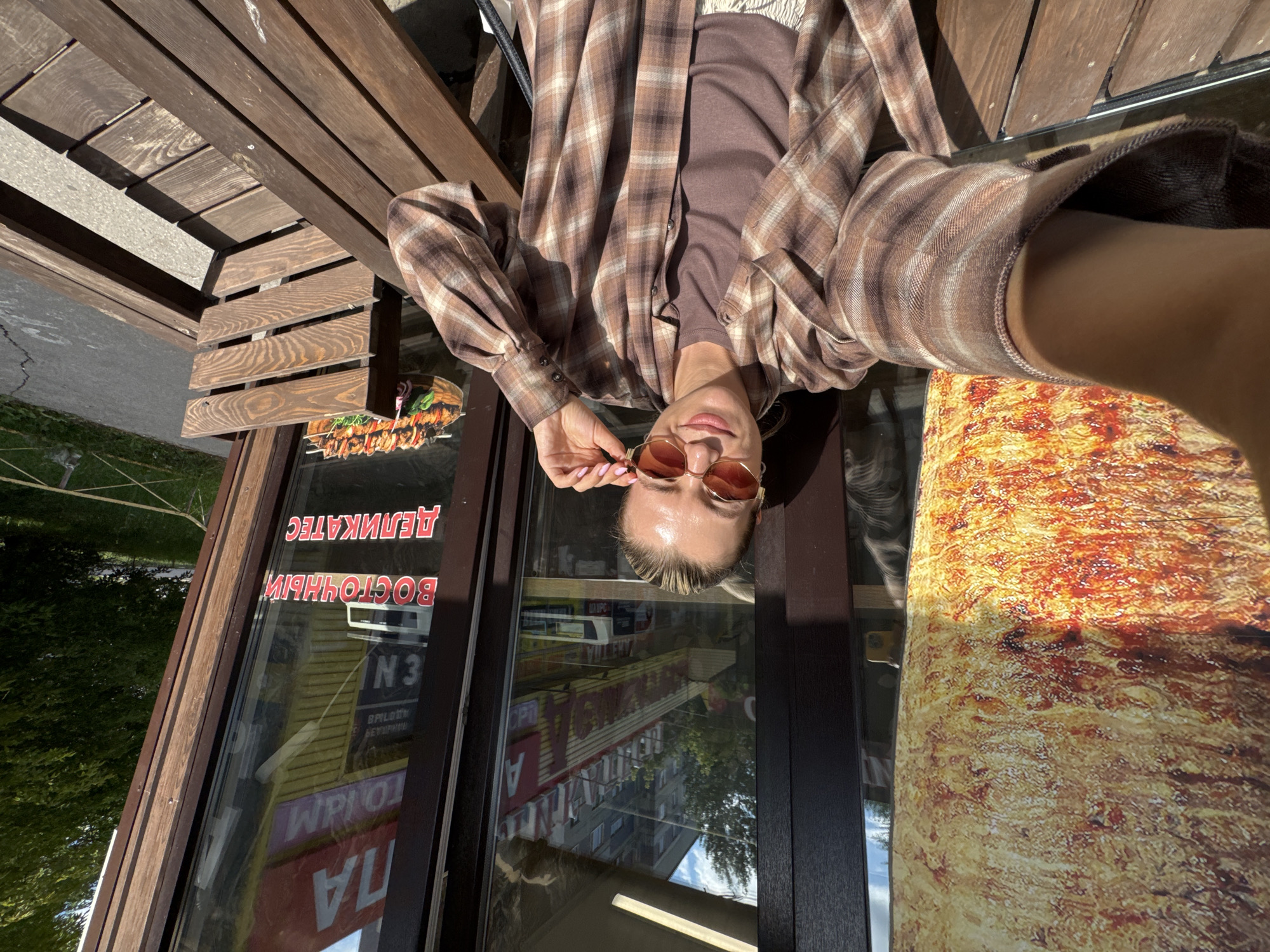 An overhead shot shows a woman sitting on a wooden bench outdoors in front of a building with a large window. The woman is wearing a brown and white plaid shirt, a brown t-shirt, and white pants. She is holding sunglasses in her right hand and is looking at the camera. A large image of stacked meat, likely for shawarma, is visible on the lower left side of the frame. Text in Russian on the building window reads "ВОСТОЧНЫЕ ДЕЛИКАТЕС." The setting appears to be a street with trees and a sidewalk visible in the background.