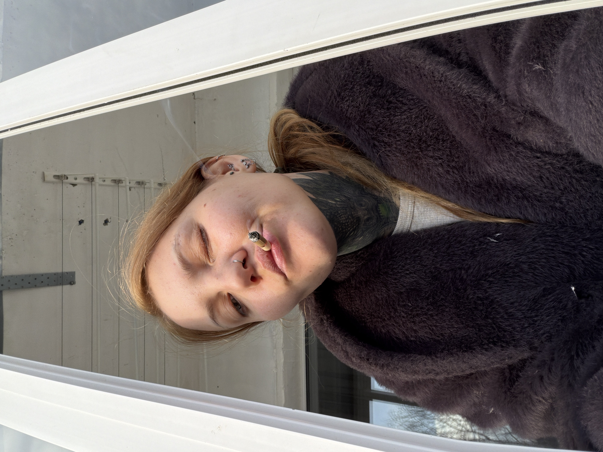 A person with fair skin, light brown hair, and visible tattoos on their neck and multiple ear piercings is holding a lit cigarette in their mouth. They are wearing a dark, furry coat or jacket. The person is positioned near a white window frame, with a view of an exterior wall visible behind them. The location is Chaikovsky, Russia.