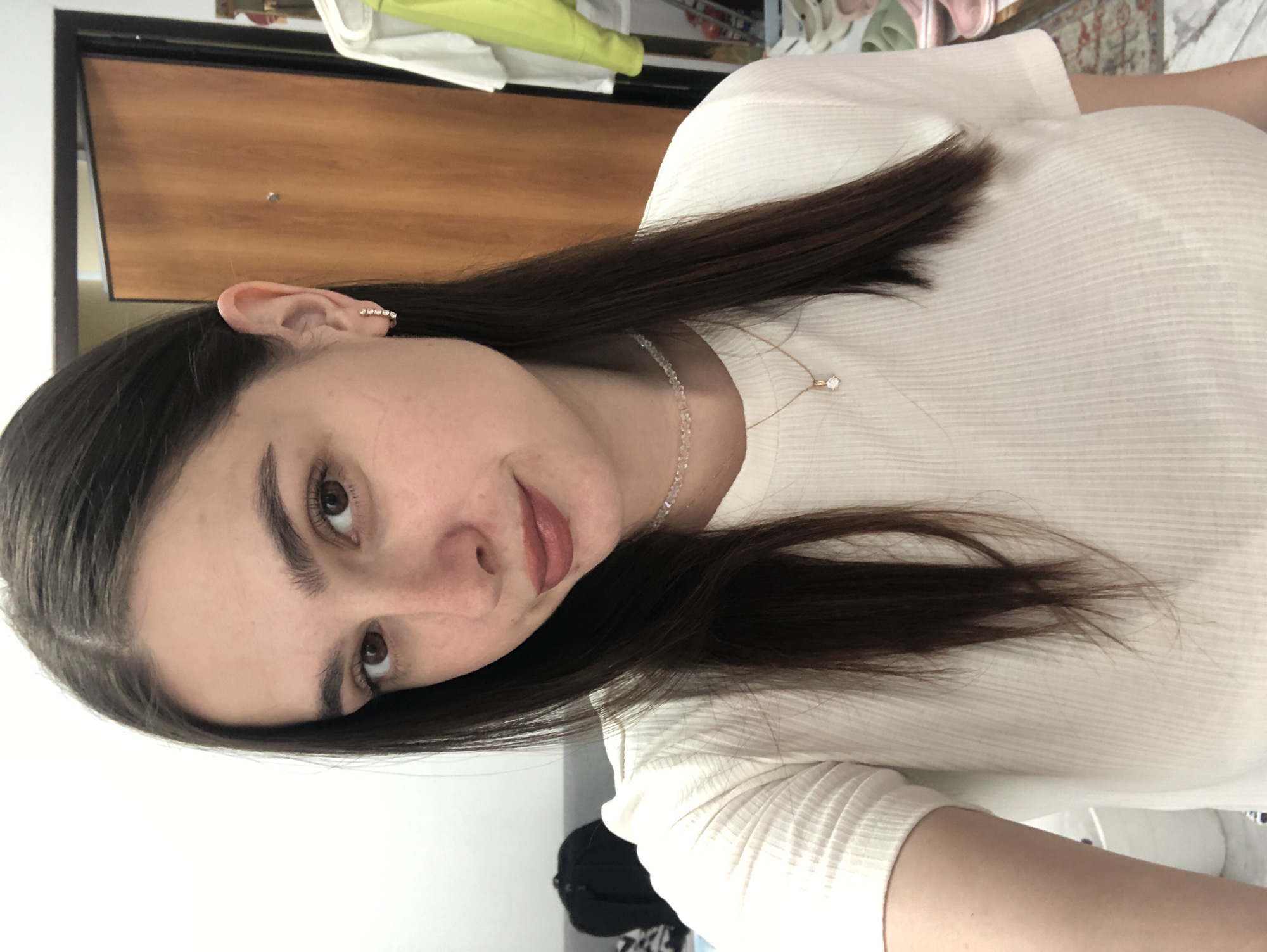 The image shows a young woman with long, dark hair, wearing a white, ribbed shirt and a delicate necklace with a small pendant. She is positioned at a slight angle, with her head tilted. Her left ear is visible and adorned with a delicate, studded earring. Her facial features, including her brown eyes and lips, are visible. The background includes a wooden door and a white wall, suggesting an indoor setting. The lighting appears to be from an overhead source, casting soft shadows. The overall impression is a casual, close-up portrait of the woman. No specific activity is discernible, and the image appears to be a still photograph. The provided location context of Chaikovsky, Russia, does not offer any visual cues within the image itself.