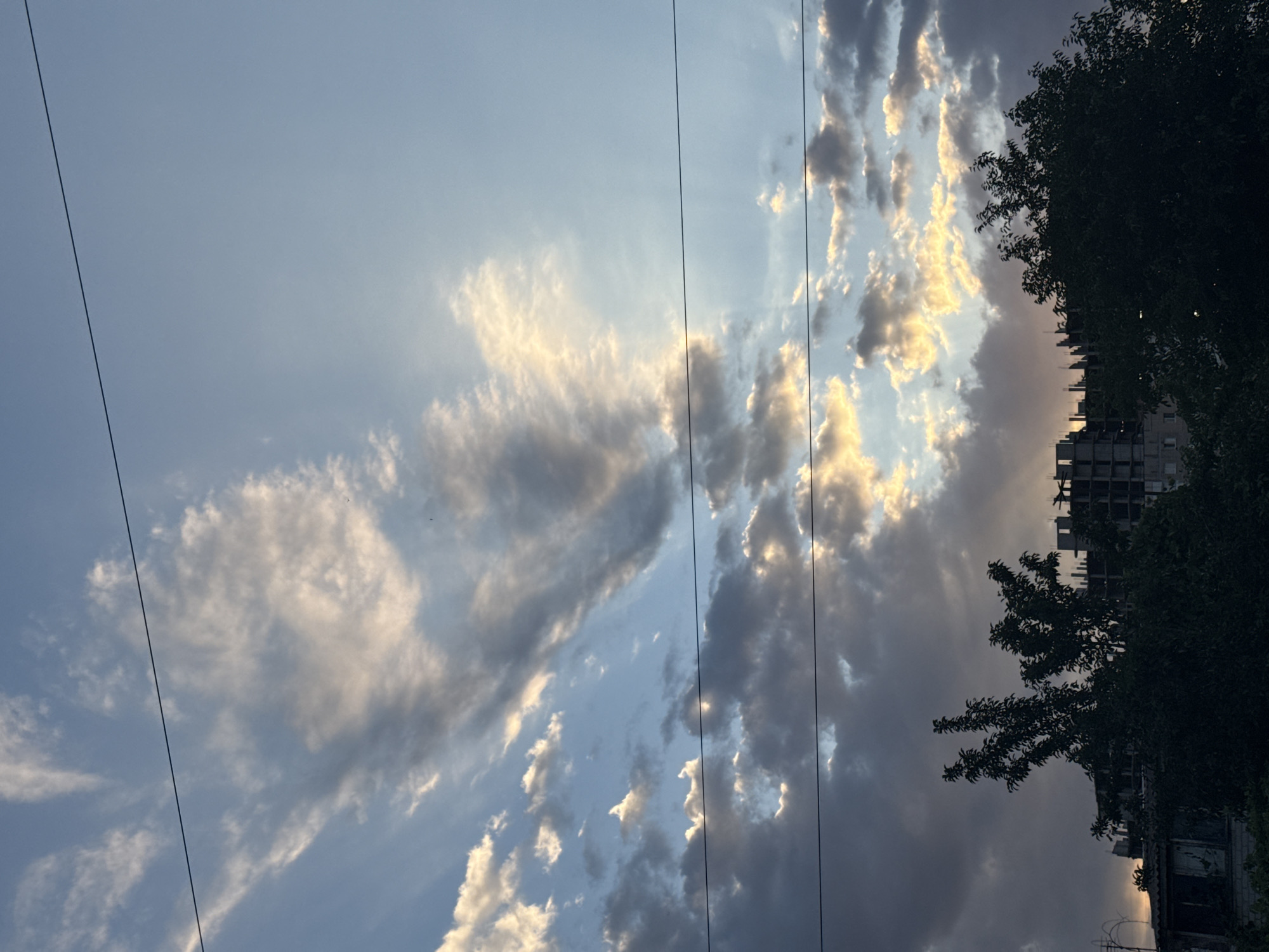The media file is a vertical, eye-level, medium shot of a cloudy sky during what appears to be sunset. The sun's rays are visible, illuminating the clouds with golden light, creating a dramatic contrast with the darker, shadowed parts of the sky. Several thin, dark electrical wires stretch across the frame, disrupting the natural beauty of the sky. On the right side of the image, a silhouetted tree with dense foliage partially obscures an unfinished or abandoned building. The building's architecture suggests a modern, possibly urban, setting. The overall mood is serene yet slightly melancholic due to the presence of the power lines and the unfinished building. The time of day is evident from the warm, directional light of the setting sun. No text is visible in the image.