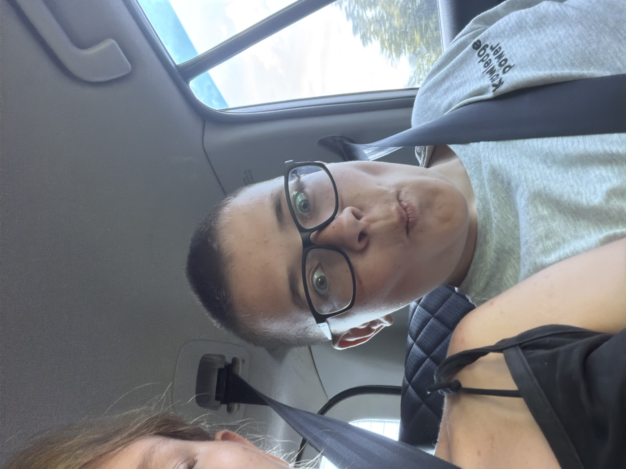 Two people, one an adult and one a child, are in the back seat of a car. The adult, on the left, has blonde hair and is wearing a black top. The child, on the right, is wearing glasses and a grey sweatshirt. The child is making a funny face. The car appears to be in motion, as indicated by the blurred trees visible through the window. The environment suggests a suburban or rural setting. There is no indication of Kruglovka, Russia in the image.
