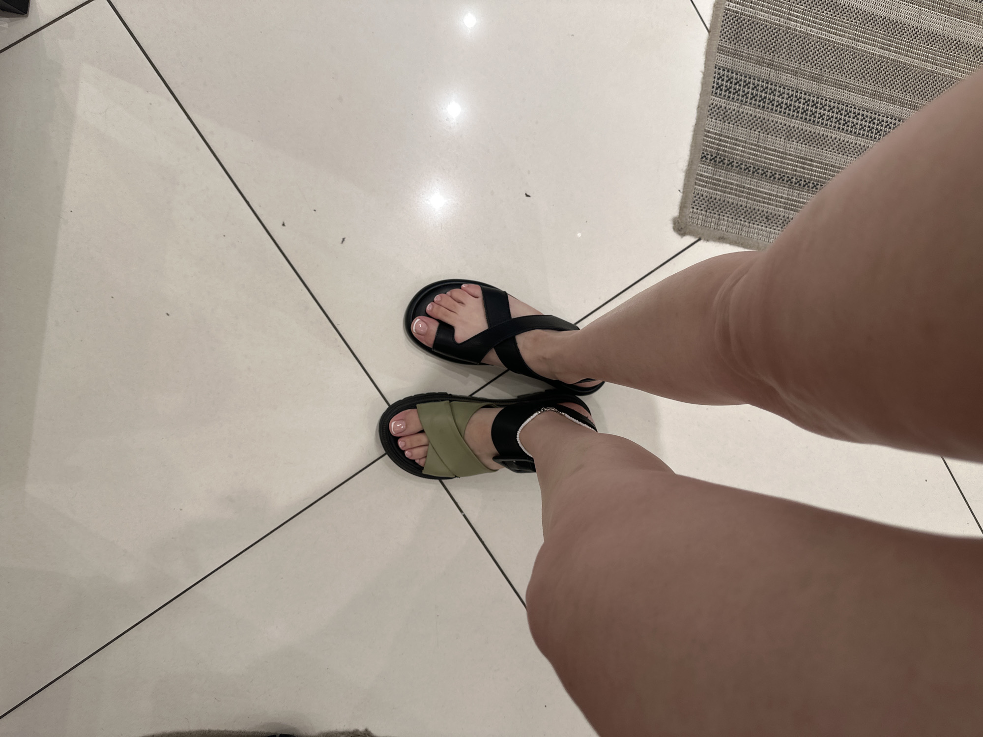 The image depicts a person standing on a light beige tiled floor with black grout lines. The person is wearing open-toed sandals; one is black with crisscrossing straps, and the other is black with an olive-green strap. The person's toes are painted with a light pink nail polish. A small portion of a gray rug is visible in the upper right of the image and a smaller portion in the lower left. The image provides no information that suggests the location is in Perm, Russia.