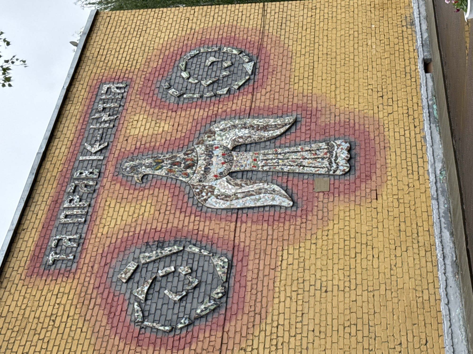 A low-angle, medium shot depicts a section of the exterior wall of a building in Chaikovsky, Russia. The wall is constructed from small, rectangular yellow bricks. Vertically oriented on the left side, mosaic lettering spells out "CHAIKOVSKY" in Russian. A mosaic eagle with outstretched wings dominates the center of the image, positioned between two circular mosaic designs. The mosaic elements are primarily comprised of small, fragmented pieces of tile. The area surrounding the mosaic designs has a reddish-brown hue, contrasting with the yellow bricks. The building's edge is visible on the right, finished with a gray material. Trees are partially visible in the upper-left corner. The lighting suggests an overcast day.