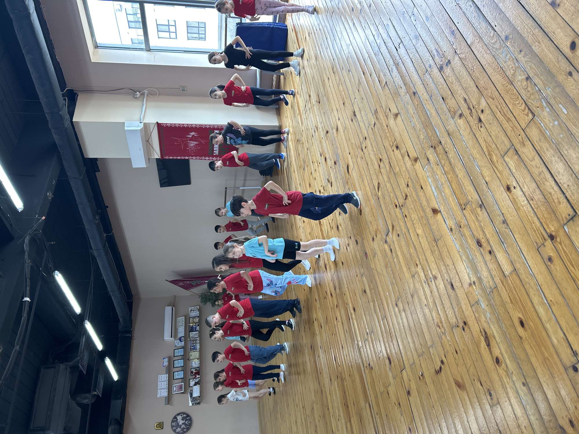 A group of approximately 15 children are in a room with a wooden floor, arranged in lines and facing the same direction, potentially practicing dance or exercise. Most of the children are wearing red shirts, with a few in different colored shirts. They all have their hands on their hips. There's a window visible in the upper left of the frame, and a red banner hanging on the wall. The room appears to be a studio or gym, based on the lighting fixtures, wooden floors, and other objects attached to the wall.
The scene is taking place indoors, and is likely located in Varna, Bulgaria, based on the description provided.