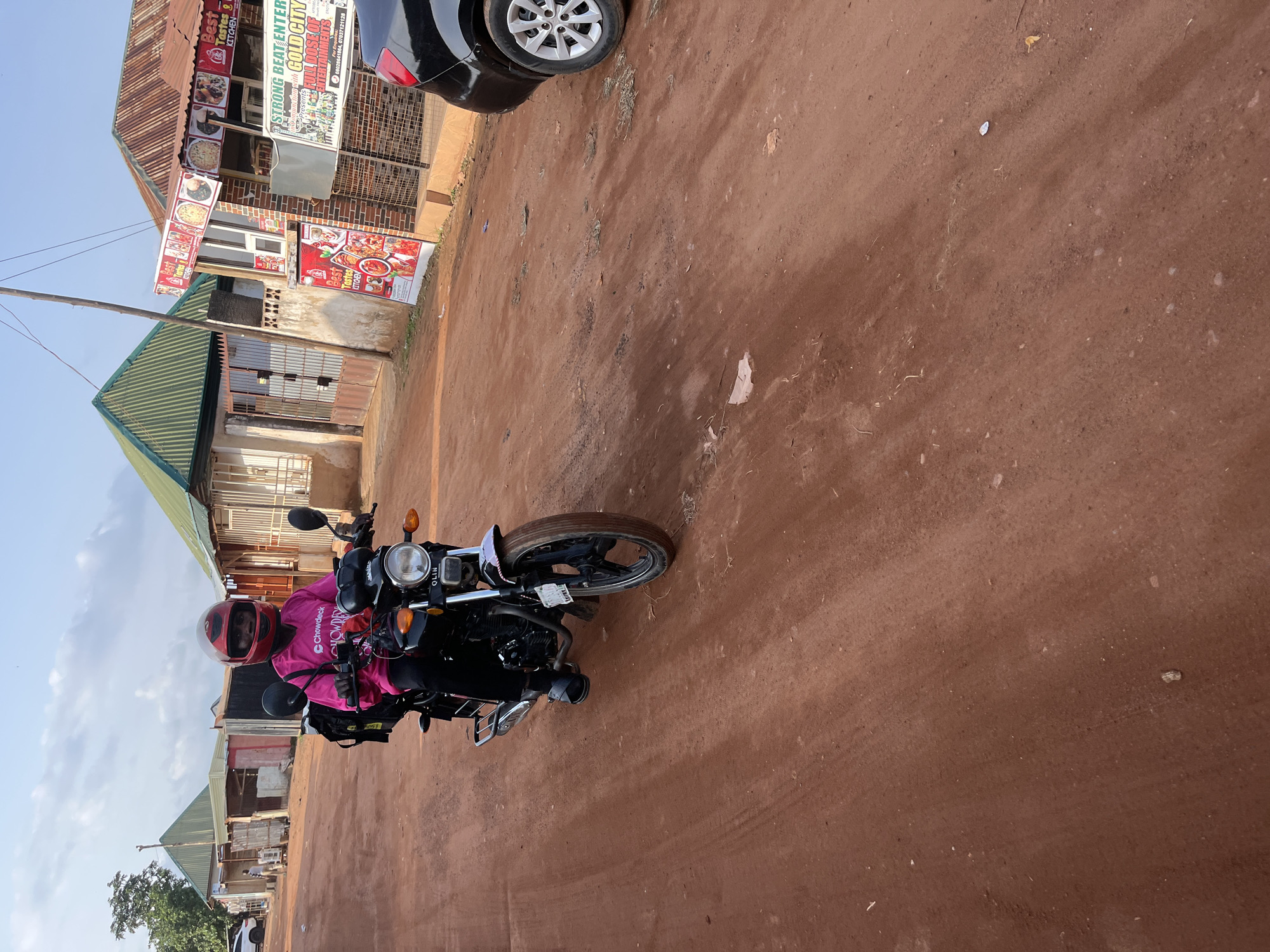 A person wearing a pink shirt, black pants, and a red helmet rides a black motorcycle on a dirt road in Abuja, Nigeria. The person is looking towards the camera and appears to be a delivery driver, indicated by the black bag on the back of the motorcycle with the "Chowdeck" logo visible. The motorcycle has a "OLIN" logo on the front. In the background, there are several buildings with corrugated metal roofs, some with storefronts and signage for businesses. One sign reads "BEST TASTES KITCHEN" and another promotes "STRONG BEAT ENTERTAINMENT with GOLD CITY." A black car is parked on the right side of the road. The ground is reddish-brown dirt with some scattered debris. The sky is overcast with clouds.