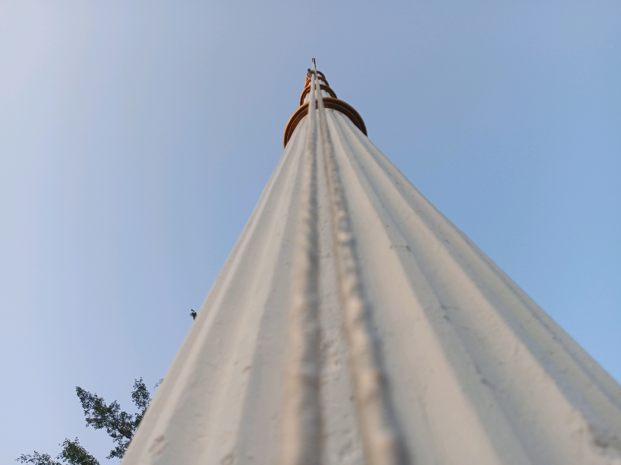 A low-angle, full shot captures a tall, white, fluted flagpole against a clear, pale blue sky. The flagpole dominates the frame, tapering upwards and seeming to pierce the sky. The texture of the flagpole is rough and marked with vertical ridges and occasional dark smudges. Towards the top, decorative gold bands encircle the pole, and a small object, possibly a flag, is visible at the very apex. In the lower left corner, the dark green foliage of trees peeks into the frame, providing a contrast to the starkness of the pole and sky. The scene is devoid of people and action, presenting a static and architectural view. The image does not provide any specific indicators of its location within Bandung City, Indonesia.