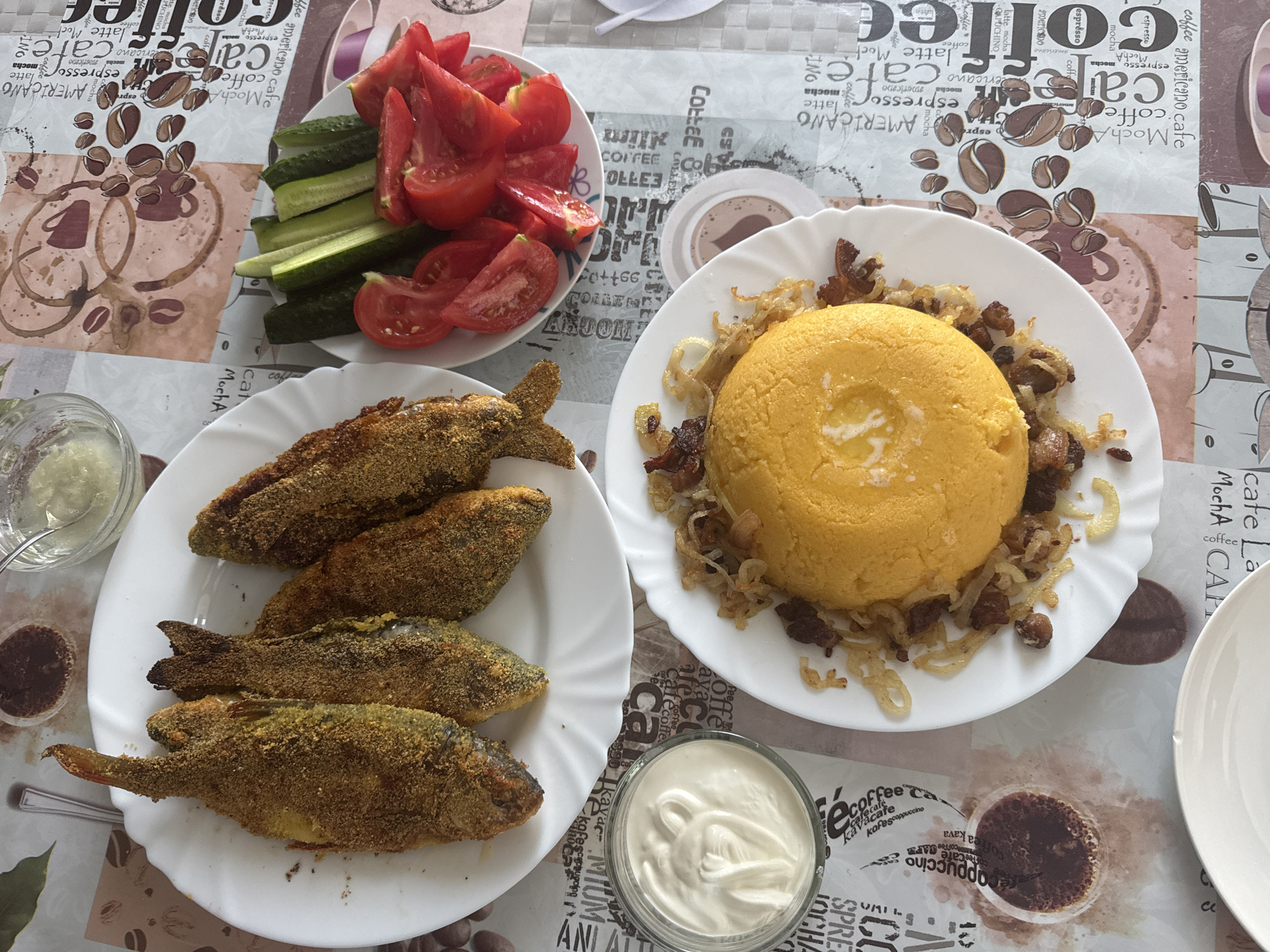 The image shows a table laden with food, likely a meal at a home or restaurant in Bălți, Moldova. In the foreground, a white plate holds a mound of polenta topped with fried onions and meat. To the left of the polenta is a small bowl of sour cream or mayonnaise. Above this, a larger white plate contains several fried fish, coated in a yellowish crust. To the right of the fish, a small white dish holds sliced tomatoes and cucumbers. A small glass of sauce is visible at the top of the image. The table is covered with a patterned tablecloth featuring coffee-related imagery. There are no people visible in the image.