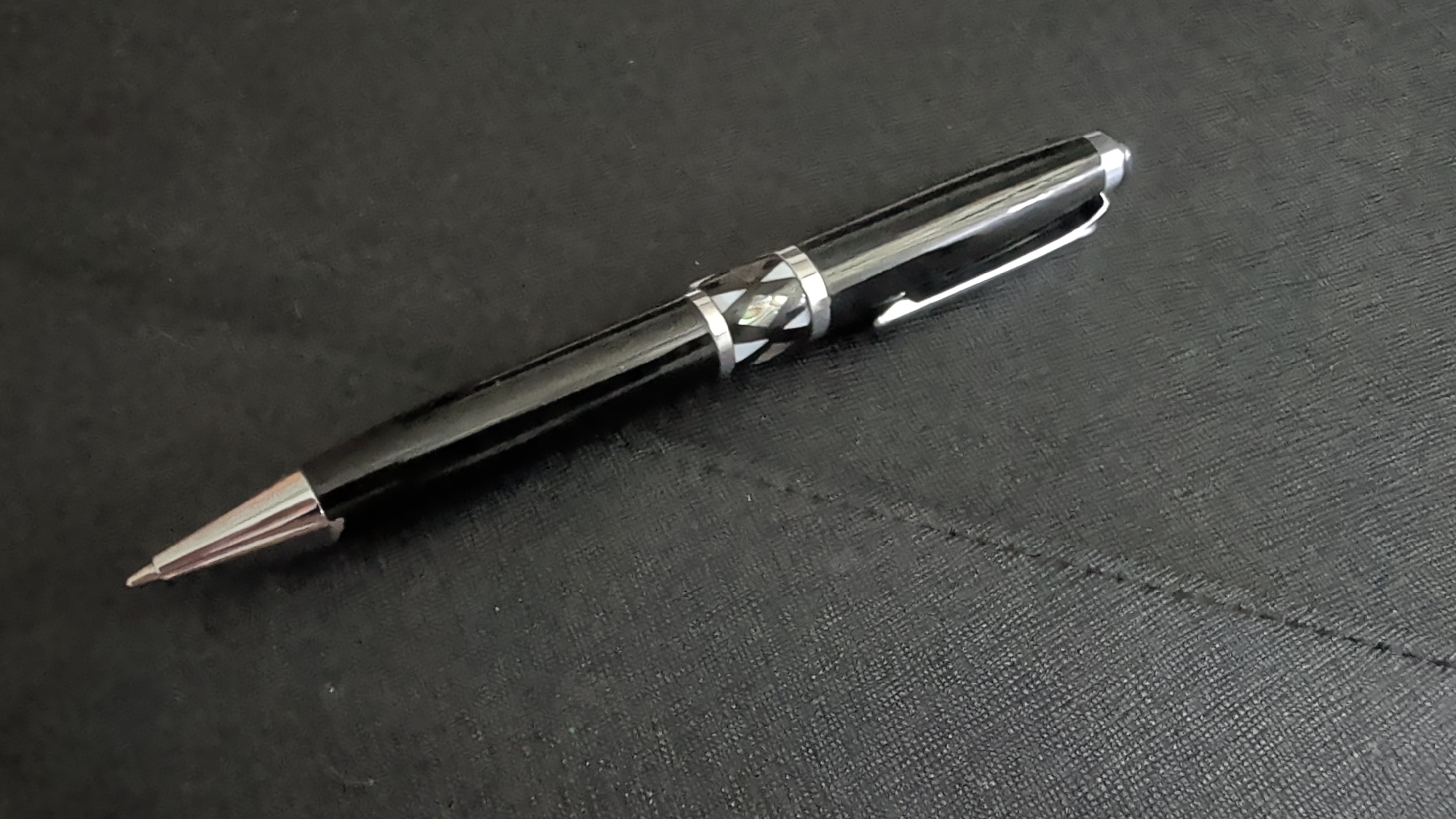 A black ballpoint pen with a silver clip and silver tip rests on a textured black surface. The pen has a decorative band around its barrel featuring a geometric pattern of alternating white and dark, possibly mother-of-pearl, inlay. The pen is positioned diagonally, with the tip pointing towards the lower left corner of the frame and the cap towards the upper right. The black surface appears to be a mat or a notebook cover with a subtle woven texture. A faint, diagonal seam or crease is visible on the surface to the right of the pen. The lighting suggests an indoor setting, likely a desk or table, with a soft, even illumination. No people or specific location cues are apparent beyond the provided context of Knittelfeld, Austria.
