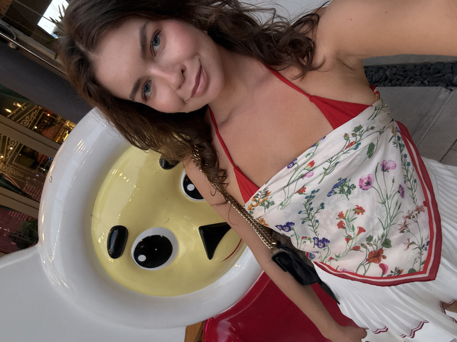 An adult female subject is depicted in a selfie-style photograph in Choeng Thale, Thailand. She has medium brown, wavy hair and fair skin, looking directly at the camera with a subtle smile. She wears a red halter-style top, a white pleated skirt with red trim, and a white scarf featuring a multi-colored floral pattern draped diagonally over her torso. A black shoulder bag with a gold chain strap is visible over her right shoulder. Positioned to her left and slightly behind her is a large, glossy, cartoon-like statue. This statue features a white rounded head, a yellow face, black oval eyes, black eyebrows, and a small black triangular nose/mouth detail, with a red lower body section. The background reveals blurred reflections from a glass-fronted building exhibiting ornate, possibly metallic, architectural or decorative details, indicative of a commercial or public complex. A dark, textured ground surface is visible on the far right.