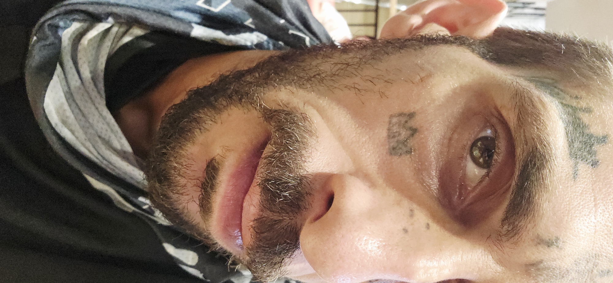 A close-up, horizontally-oriented portrait of a person with fair skin, a dark beard and mustache, and dark hair. The person has tattoos on their face, including a "B" or "13" near their right eyebrow and a star-shaped tattoo above their left eyebrow. The person is wearing a dark garment with a patterned head covering. The background is blurry and indistinct. There is no indication of the location being Bắc Nha Trang, Vietnam.