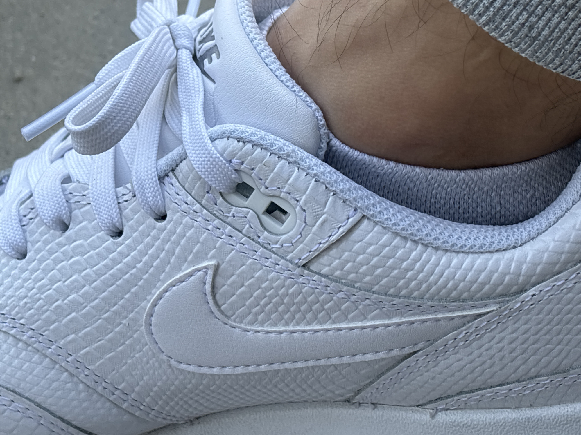 A close-up photograph shows the upper portion of a white sneaker being worn. The shoe features a textured, possibly snakeskin-like pattern, across its surface, including the iconic Nike swoosh logo. The laces are white and tied. A gray sock is visible around the ankle of the wearer. The setting is not discernible, and there is no indication that it is specifically in Plovdiv, Bulgaria. There are no people fully visible, only a partial view of the ankle and leg of the individual wearing the shoe.