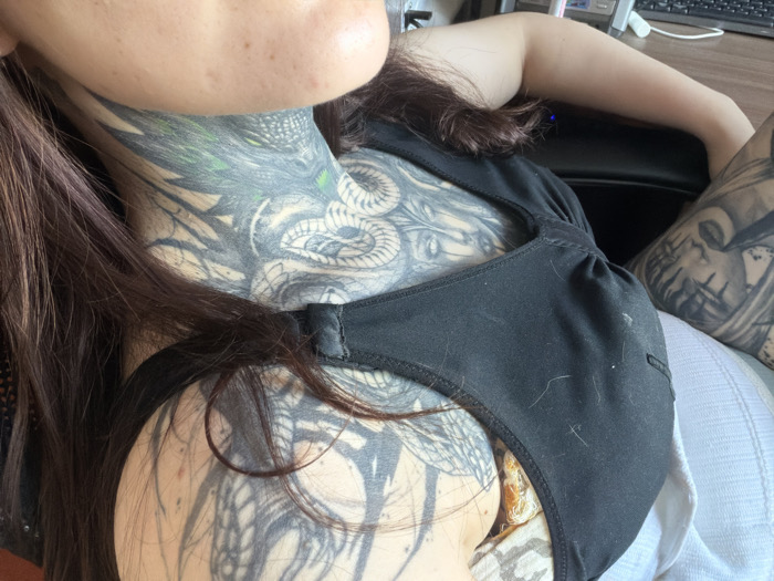 A woman is seated, wearing a black tank top with visible tattoos on her neck, chest, and arms. The neck tattoo features a serpentine design. Dark brown hair drapes across her shoulder and upper chest. The background includes a computer keyboard and other office equipment, suggesting an indoor setting. A white cloth or pillow is visible behind her right arm.