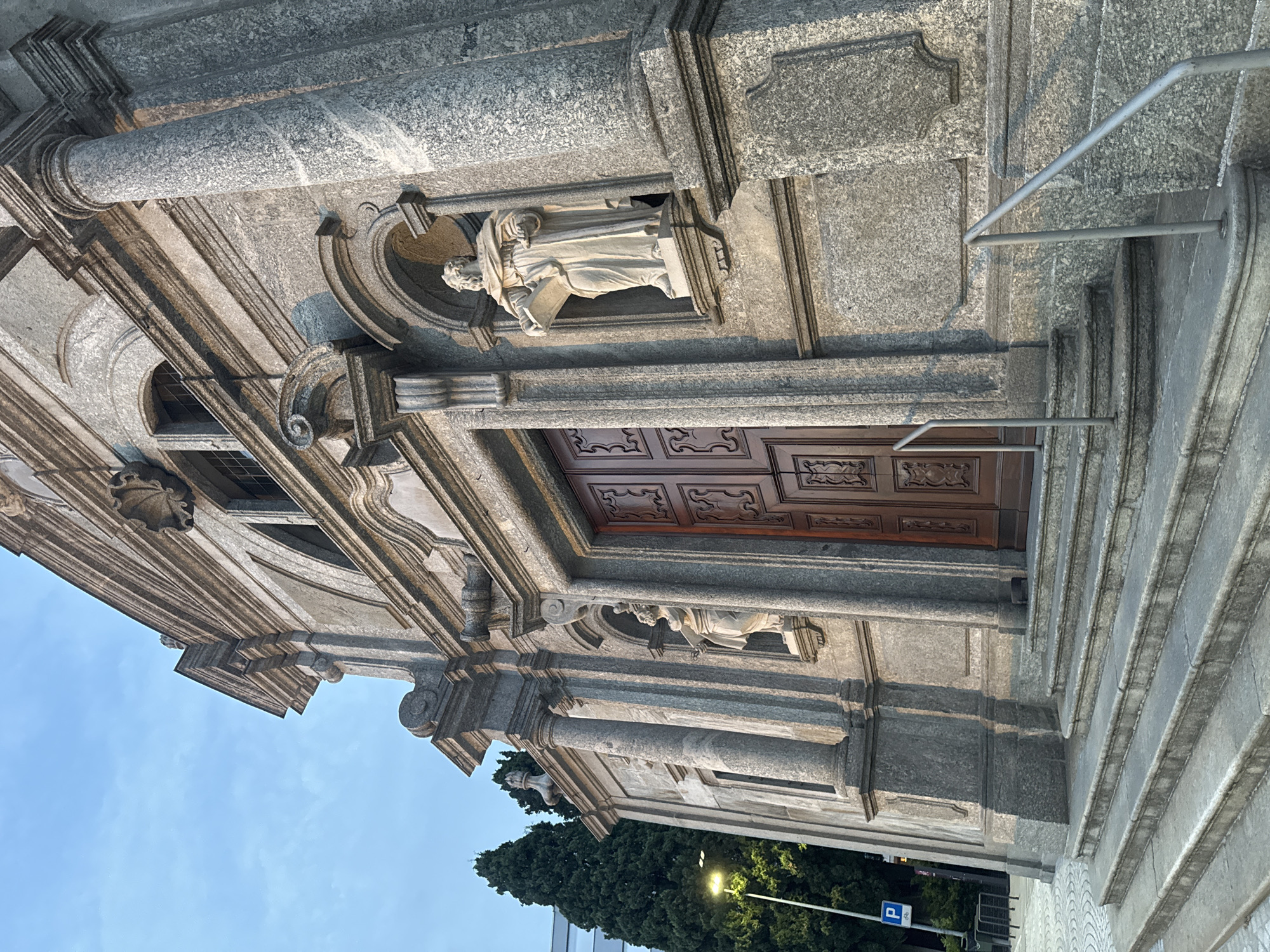 The image shows the facade of a stone church in Balerna, Switzerland, with a clear blue sky overhead. The church features grand wooden doors, flanked by columns and adorned with statues. In the foreground, stone steps lead up to the entrance, with metal railings on either side. To the left of the church, tall evergreen trees and a lamppost are visible, along with a parking sign. The lighting suggests it is late afternoon or early evening, with shadows cast across the steps and facade.