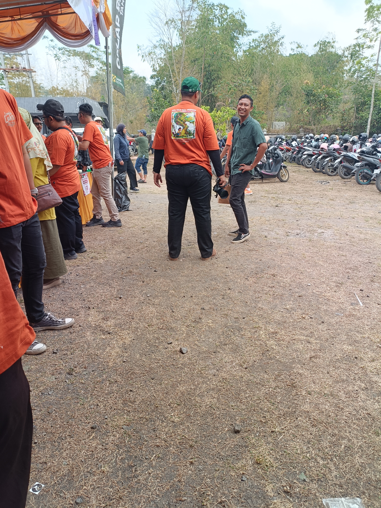 The image depicts an outdoor event likely held in Hargobinangun, Indonesia, given the context provided. The foreground is dominated by a dry, grassy, and rocky ground. In the mid-ground, a group of people are present. One man, wearing an orange t-shirt with a graphic print and a green cap, is facing away from the camera. Another man, in a green collared shirt and dark pants, is smiling and facing the camera. To the left, several individuals are seen in orange t-shirts and other casual attire. Behind the people, a row of parked motorbikes is visible. In the background, trees and a cloudy sky are present, suggesting an overcast day. A tent structure with white and orange fabric is partially visible on the far left. The overall scene suggests a community gathering or event, possibly related to motorcycling or local activities.