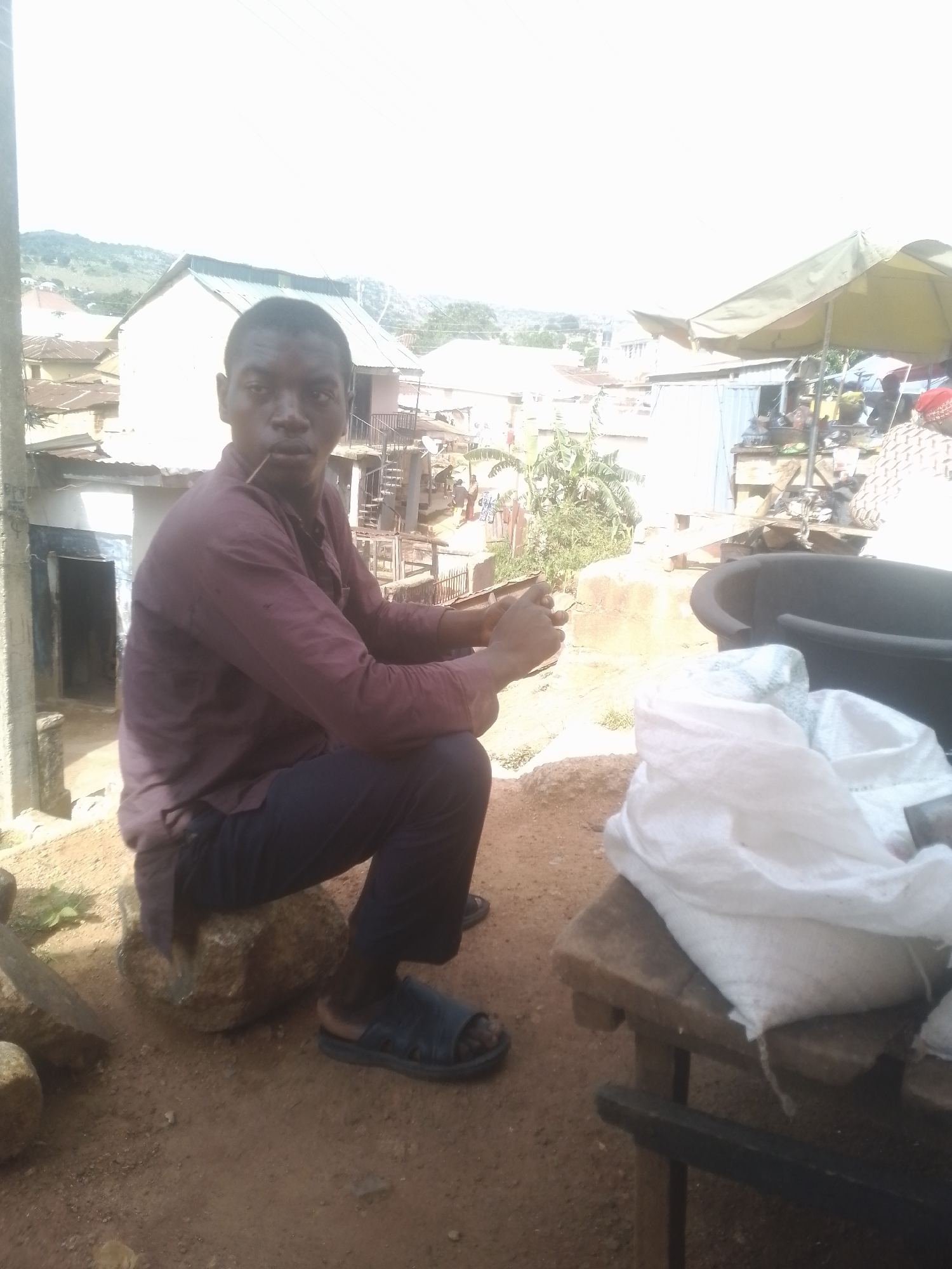 A full shot shows a young Black male sitting on a rock outside, looking into the camera. He is wearing a dark red shirt and dark pants with blue sandals. He holds a twig in his mouth and his right hand is raised to his face, holding what appears to be another twig. Behind him, a cluster of buildings with corrugated metal roofs are visible, with trees and hills in the distance. To the right, a large black tub and a white bag on a wooden table are partially visible. The ground is dirt, with some scattered rocks. The sky is bright and overcast.