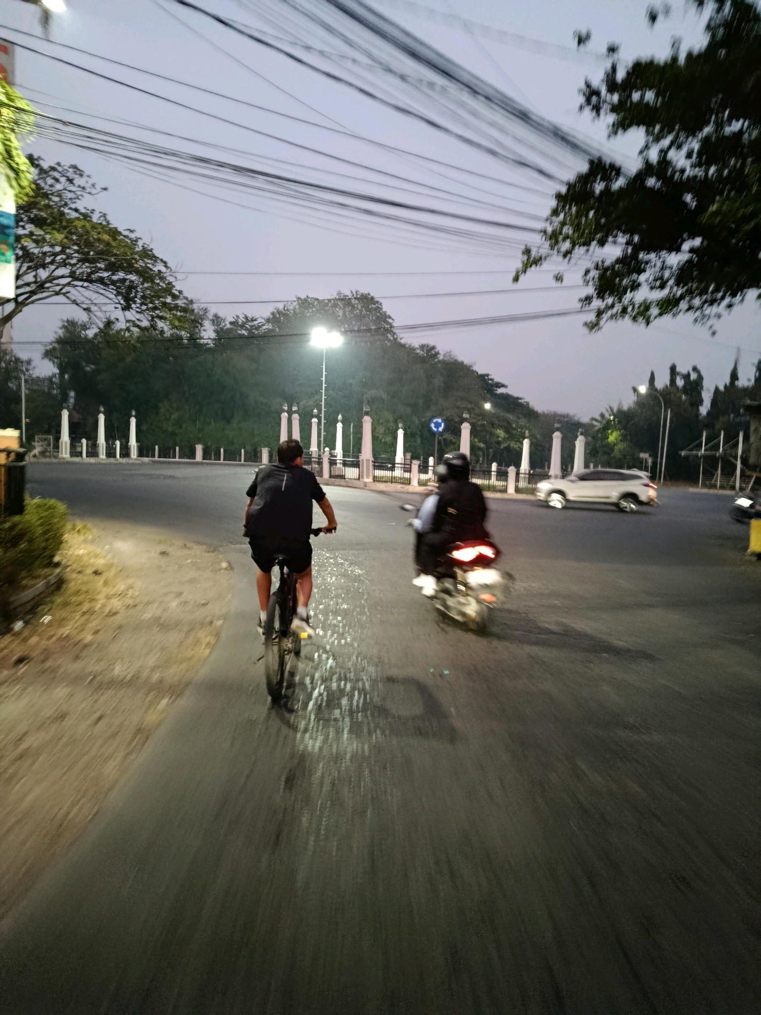 In Bekasi, Indonesia, at dusk, two individuals are captured in motion on a street. The foreground features a cyclist, viewed from behind, pedaling a bicycle along a paved road. The cyclist wears a dark t-shirt and shorts, with a backpack on their back. To the right of the cyclist, a motorcycle carrying two individuals is also in motion. The motorcycle rider wears a helmet and a backpack, while the passenger is similarly attired. Both vehicles are moving away from the camera, creating motion blur.
The background reveals a street intersection with several white pillars lining the perimeter. Trees are visible behind the pillars, and utility poles with numerous wires stretch across the sky. A white SUV is in the process of turning at the intersection, with its wheels blurred by motion. Streetlights illuminate the scene, casting a soft glow. The overall environment suggests a suburban or semi-urban area in Bekasi during the evening transition from daylight to darkness.