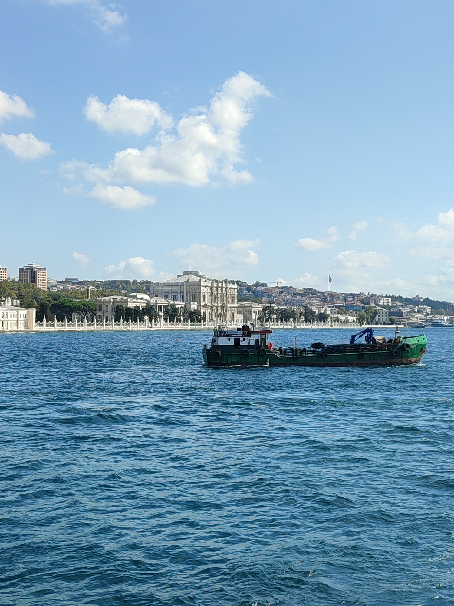 A wide-angle, medium shot shows a body of water in the foreground and a city skyline in the background under a partly cloudy sky. The water is a deep blue with ripples on its surface, indicating a slight breeze. A green barge, appearing to be a workboat or cargo vessel, is positioned in the middle right of the frame, moving from left to right. The barge has a prominent crane on its stern. In the background, a grand white building with ornate architecture, likely a palace or historical government building, dominates the skyline, flanked by other structures and trees. The city appears to be situated on a gentle slope leading down to the water. The overall impression is a serene maritime scene with an impressive urban backdrop. The location is the Bosphorus Strait in Istanbul, Turkey, with the Dolmabahçe Palace, located in the Beşiktaş district (formerly part of Beyoğlu's administrative region), being the most prominent structure visible.