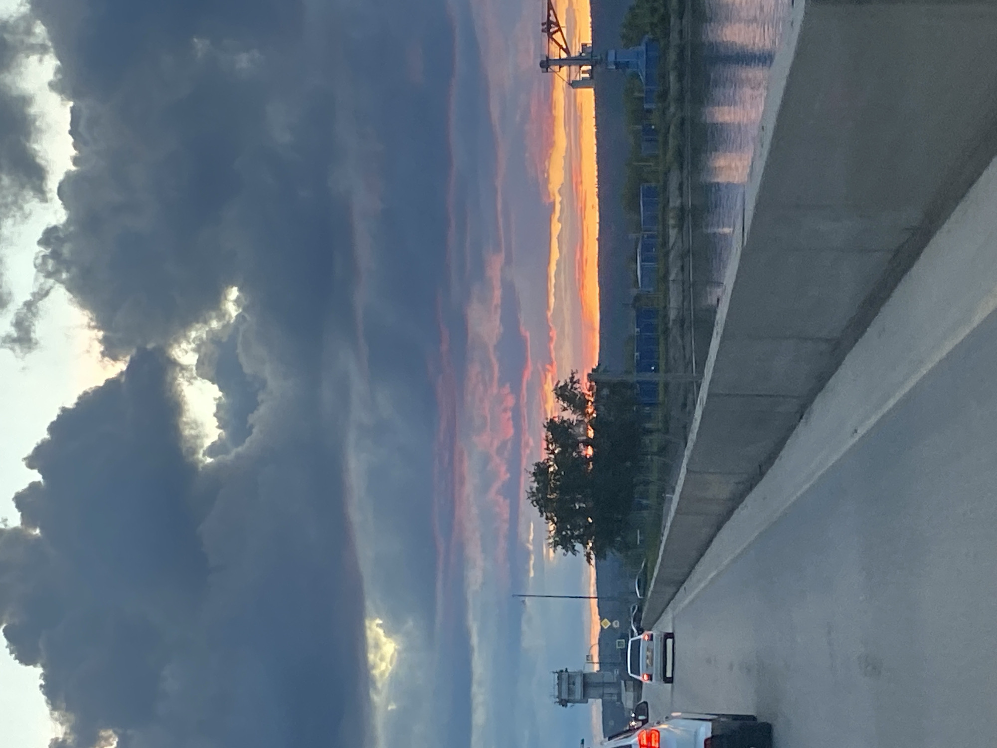 The image depicts a view from an elevated road, possibly a bridge or overpass, in the city of Chaikovsky, Russia. The sky is dominated by dark, heavy clouds on the left side, contrasting with a vibrant sunset on the right, featuring orange and pink hues. A body of water, reflecting the sunset colors, is visible beyond a line of trees and structures. The foreground shows a section of the road with vehicular traffic, including white cars and other vehicles with illuminated tail lights. A concrete barrier runs along the side of the road. A lattice structure can be seen in the distance against the horizon.