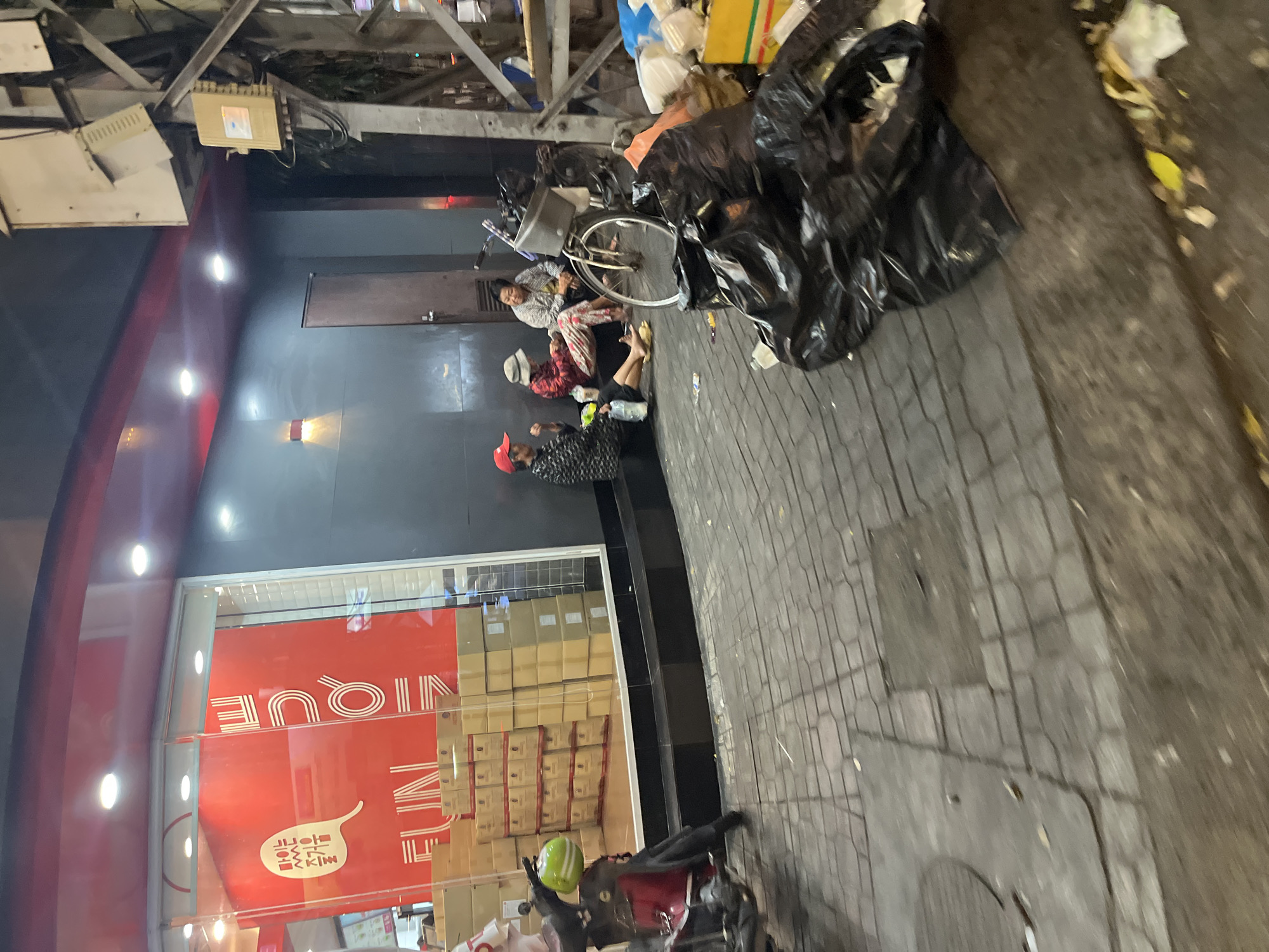 The image shows an exterior street scene, apparently in Nha Trang Ward, Vietnam, as evidenced by the signage on the storefront. Three individuals are visible on the sidewalk in front of a building with a red storefront that reads “UNIQUE” in English and a speech bubble. These individuals appear to be unhoused. The individual furthest from the camera is lying down while the other two individuals are sitting upright. A bicycle is present next to one of the individuals. There are black trash bags and other litter on the sidewalk. A scooter is parked on the sidewalk near the storefront window. The sidewalk is paved with brick tiles. There is a building structure with metal supports and utility boxes overhead.