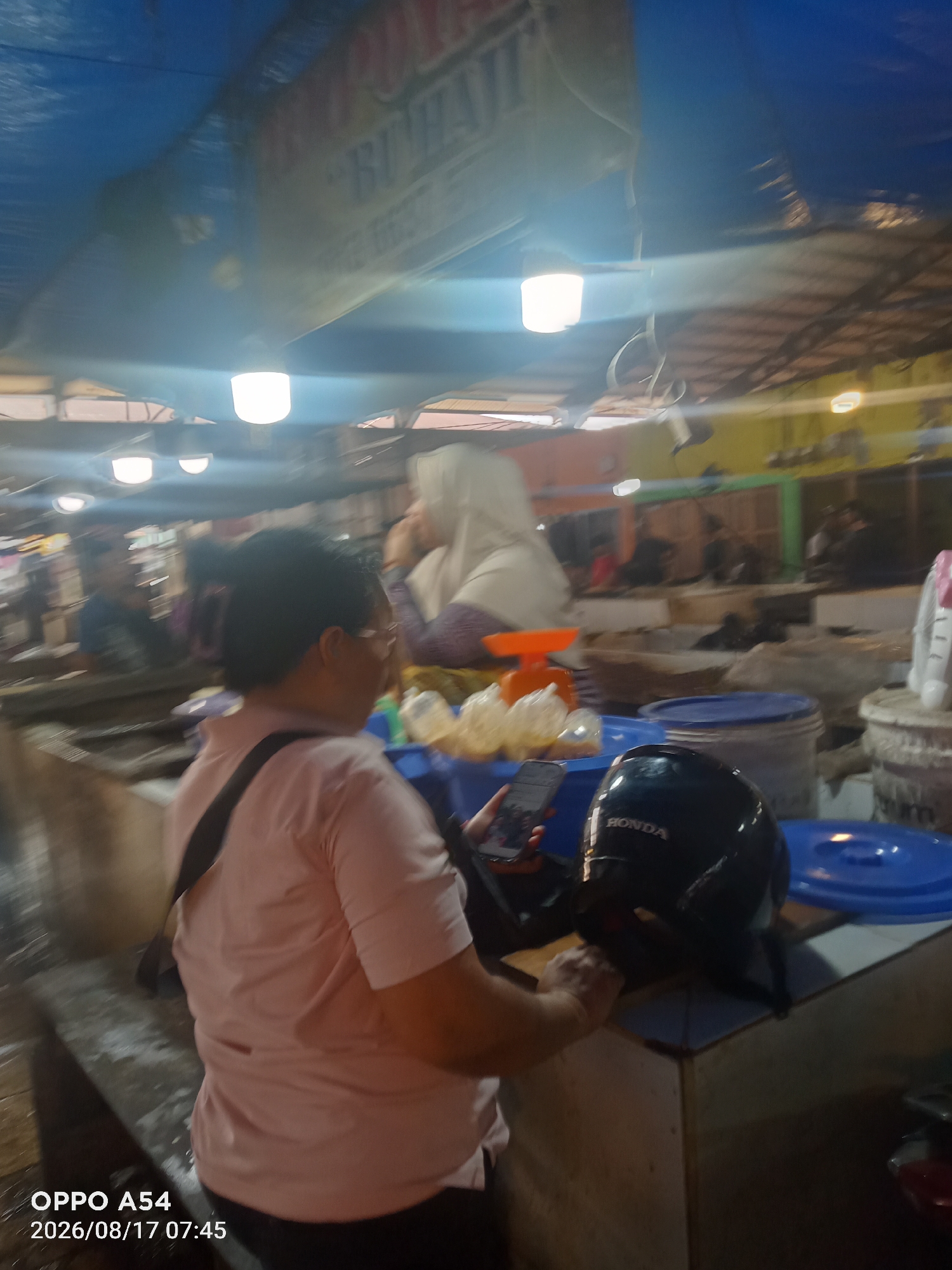 The image depicts a marketplace scene, possibly in Jambi, Indonesia. In the foreground, a person wearing a light pink shirt and glasses is holding a smartphone and looking down at it. They have dark hair tied back. Beside them, a black motorcycle helmet with the word "HONDA" is placed on a counter. In the background, other individuals are visible, including a person wearing a white hijab. Various market stalls with what appear to be goods for sale are also present. The environment seems to be covered by a blue tarp, and several lights are illuminating the area. The overall lighting suggests it is daytime, but possibly indoors or under a shaded structure. A timestamp in the lower left corner reads "OPPO A54 2026/08/17 07:45".