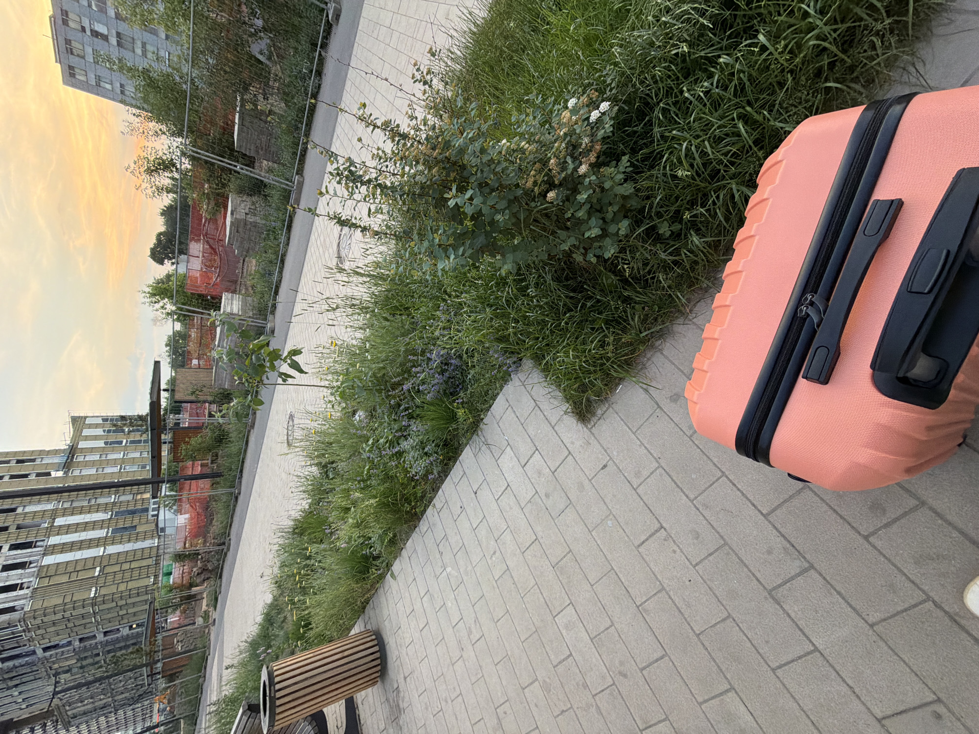A bright coral suitcase with black trim rests on a light gray paver sidewalk in Moscow, Russia. The suitcase is angled towards the lower right of the frame. Behind the sidewalk, a strip of green grass and wild vegetation runs alongside a paved walkway. The walkway is bordered by a chain-link fence with orange construction netting behind it. Beyond the fence, several large, unfinished buildings are under construction, with scaffolding and exposed structural elements visible. The sky above is a pale yellow and orange, suggesting either sunrise or sunset. A cylindrical wooden trash receptacle is visible to the left of the suitcase, also on the sidewalk.