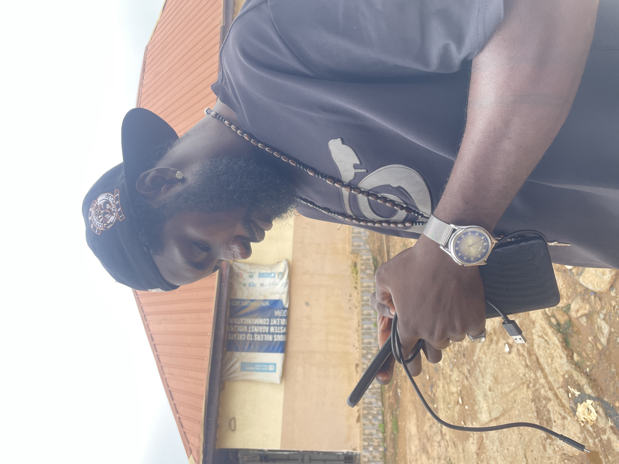 In Kurunduma, Nigeria, a man with dark skin and a beard wears a black baseball cap and a dark t-shirt. He is holding a smartphone and a charging cable. A beaded necklace hangs around his neck, and he wears a wristwatch on his left wrist and a ring on his right ring finger. Behind him, a building with a red-tiled roof and light-colored walls is visible, with a banner hanging on the facade. The ground in front of the building appears to be unpaved and somewhat muddy. The sky is overcast.