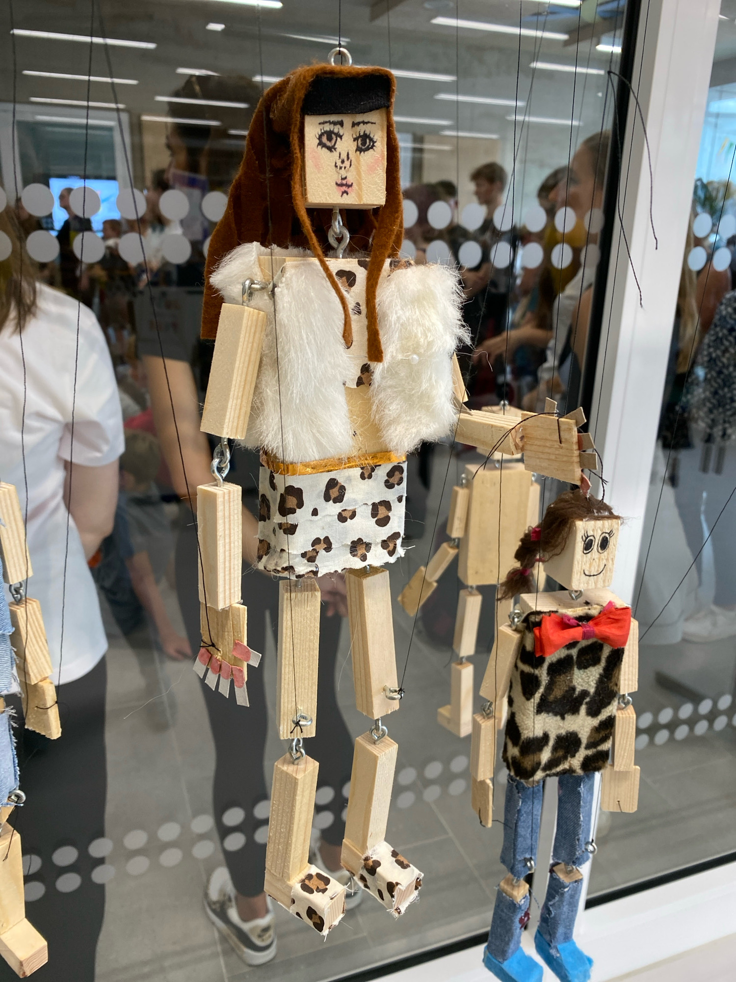 The image shows a close-up of several handmade wooden puppets hanging by strings. The puppets are designed with block-shaped wooden bodies, and some have fabric clothing and embellishments. The puppet in the foreground on the right is wearing a leopard-print textured fabric top with a red bow tie and blue denim-like pants. Its feet are bright blue. To the left and behind it is a larger puppet, also made of wood, with a white furry vest and a skirt or lower body section with a leopard print. It has a brown felt hat and hood. In the background, blurred figures of people are visible, suggesting an indoor setting, possibly a public space or exhibition. Reflections on the glass surface in front of the puppets show more of the environment and people. The overall impression is of a display of craft or art. The image does not provide any information that allows for its location to be determined as Horhausen (Westerwald), Germany.