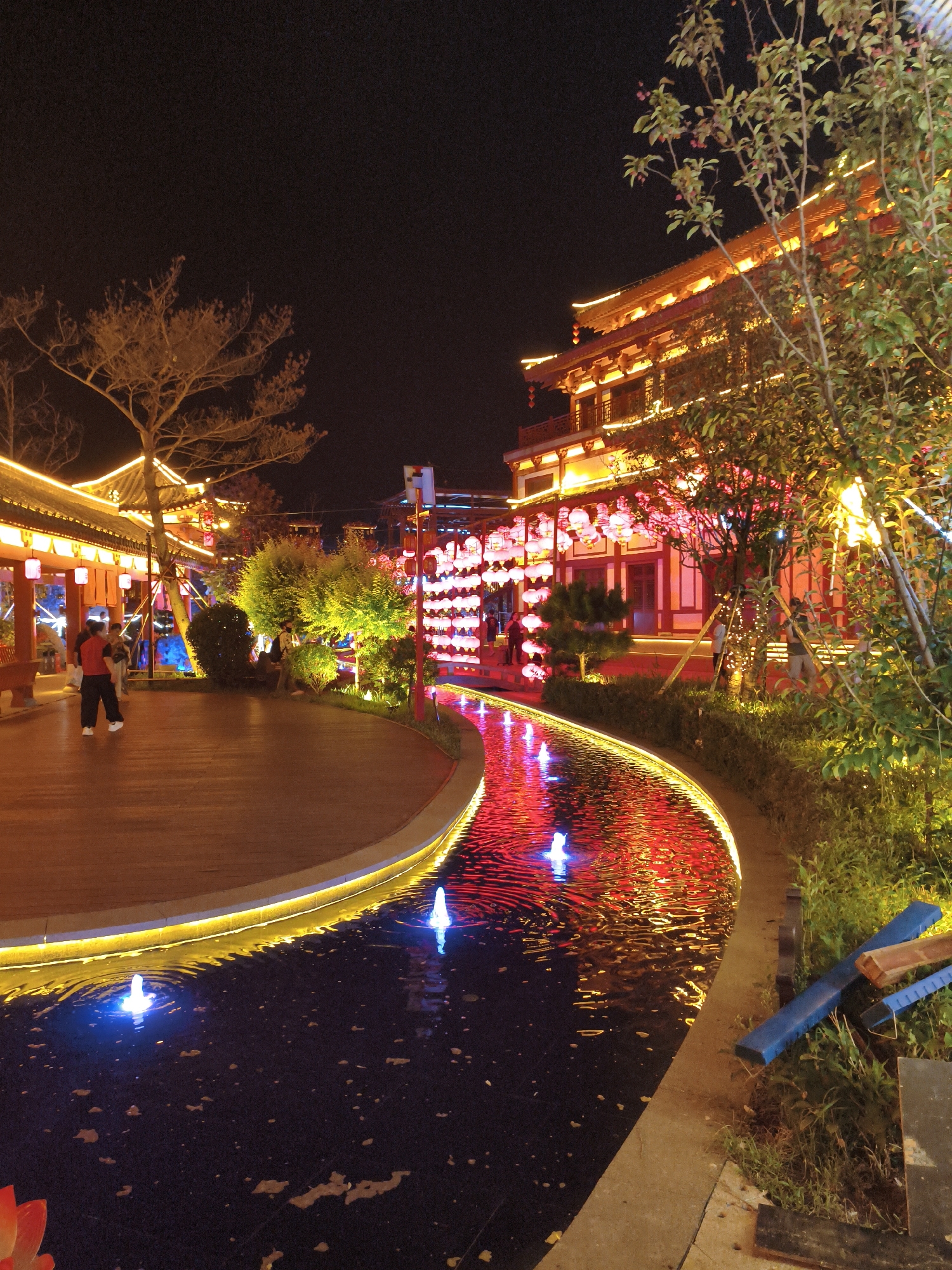 At night in Ying'an, China, a scenic area features traditional Chinese architecture with red and gold embellishments, illuminated by numerous lanterns and string lights. A curved, dark water feature with small fountains is bordered by a pathway lined with yellow lights. On the left, a wooden platform is visible with at least two individuals. One person, wearing a red shirt and dark pants, is standing on the platform. Further back, a building with tiered roofs and glowing windows is prominent. Green trees and bushes, some adorned with lights, are situated throughout the scene, with a larger tree on the left and a cluster of trees and bushes on the right. The overall ambiance is festive and illuminated.