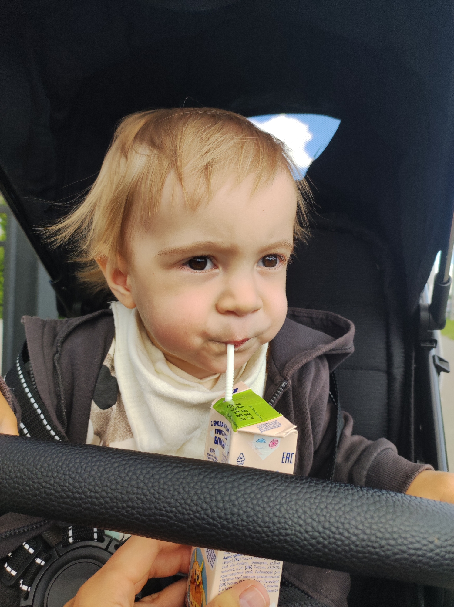 A toddler with light brown hair is sitting in a black stroller, drinking from a juice box through a straw. The juice box has a green top and white body with writing in Russian. The toddler is wearing a brown hooded jacket over a patterned shirt and white bib. The stroller has a black leather bar in front of the child. Outside the stroller, there is a glimpse of greenery and a building. Based on the Russian writing on the juice box, the location may be the city of Chaikovsky, Russia, but this cannot be confirmed from the image alone.