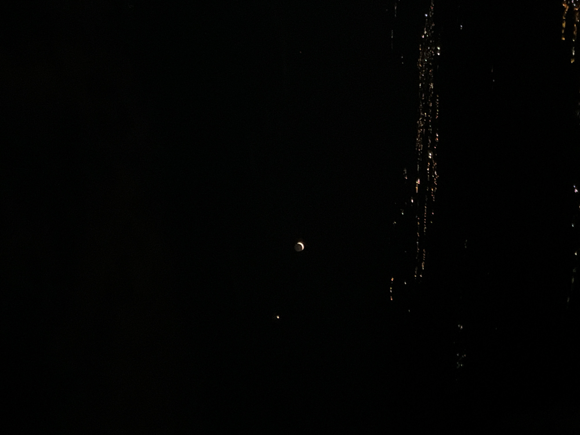 The image shows a crescent moon and a bright dot (likely a planet or star) in a dark sky. To the right of the moon and dot is a vertical band of shimmering light, likely city lights seen through a window or some reflective surface. Given the prompt's reference to Sofia, Bulgaria, it is possible this photograph was taken from an elevated position within the city, with the shimmering light representing reflections of the urban environment. The environment is largely dark, emphasizing the celestial bodies and the artificial light source.