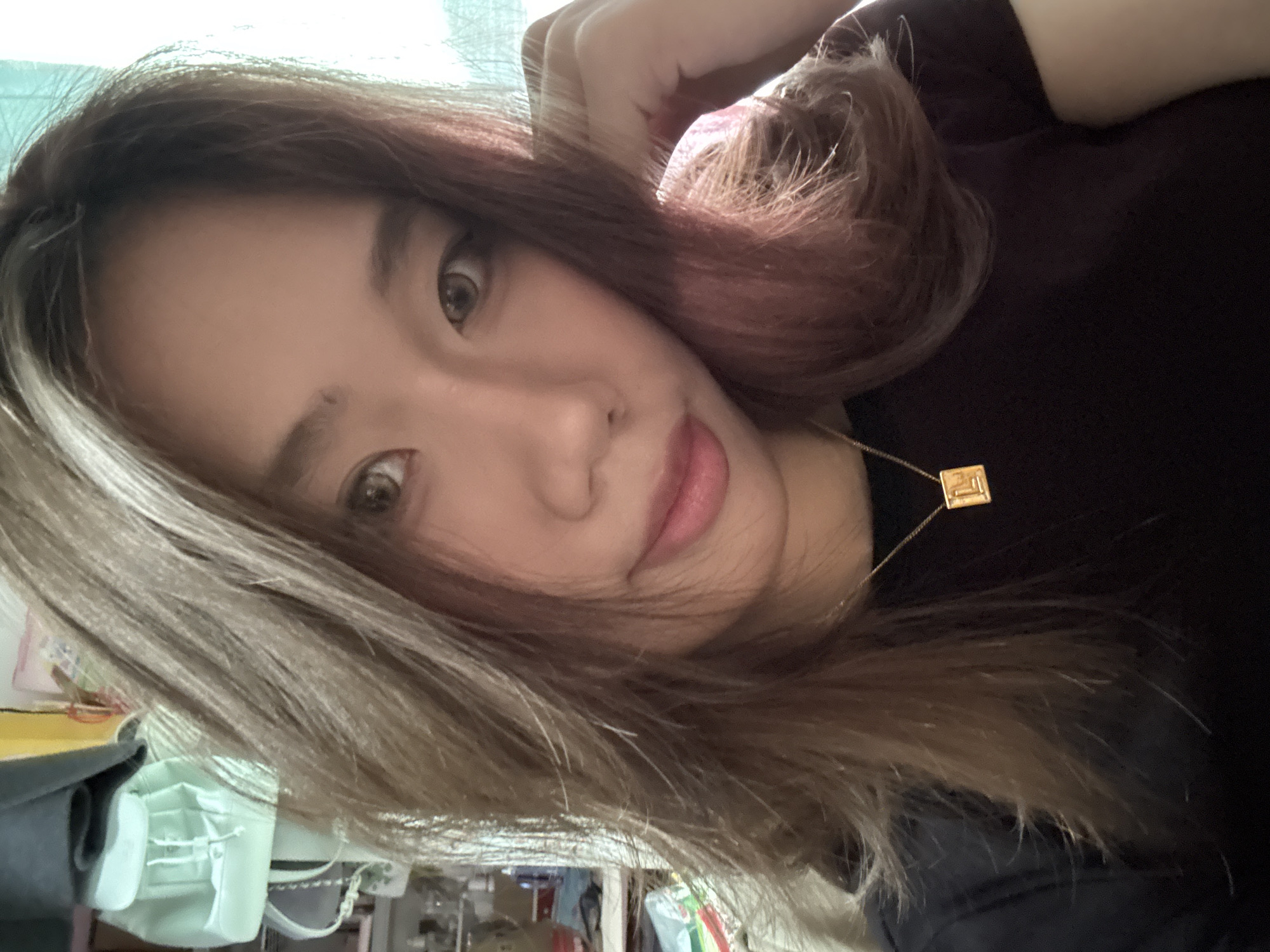 A woman with brown hair with blonde highlights and pink tones poses indoors, glancing at the camera. She is wearing a dark-colored shirt and a gold necklace with a square pendant. The background appears to be a cluttered interior with medical supplies. The specific location within Nan'an District, China, cannot be determined from the given image.