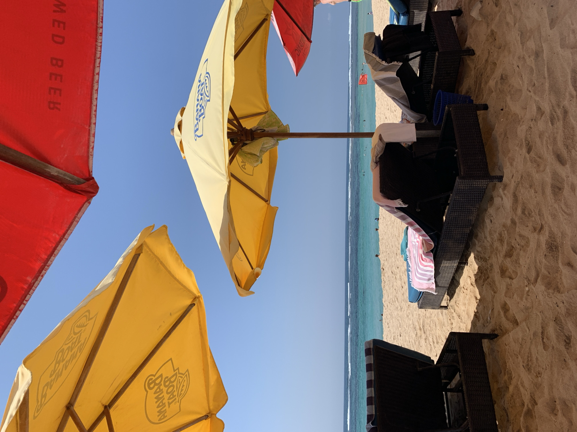 The image depicts a serene beach scene in Kuta Selatan, Indonesia, likely at a resort or public beach area. The foreground is dominated by sandy beach with several dark-colored lounge chairs, some of which are covered with towels in various colors, including pink and white stripes, and a gray and white striped towel. Two large, yellow beach umbrellas are prominently featured, one closer to the foreground and another further back. The umbrella in the foreground displays the "Banana Boat" logo. In the background, a red beach umbrella is partially visible. Beyond the sand, the turquoise waters of the ocean stretch towards the horizon, with gentle waves and a clear blue sky above. Several small figures can be seen in the water and on the beach, suggesting a day of leisure. A red flag is planted on the beach in the distance, possibly indicating a swimming advisory or a specific zone. The overall atmosphere is one of relaxation and summer vacation.