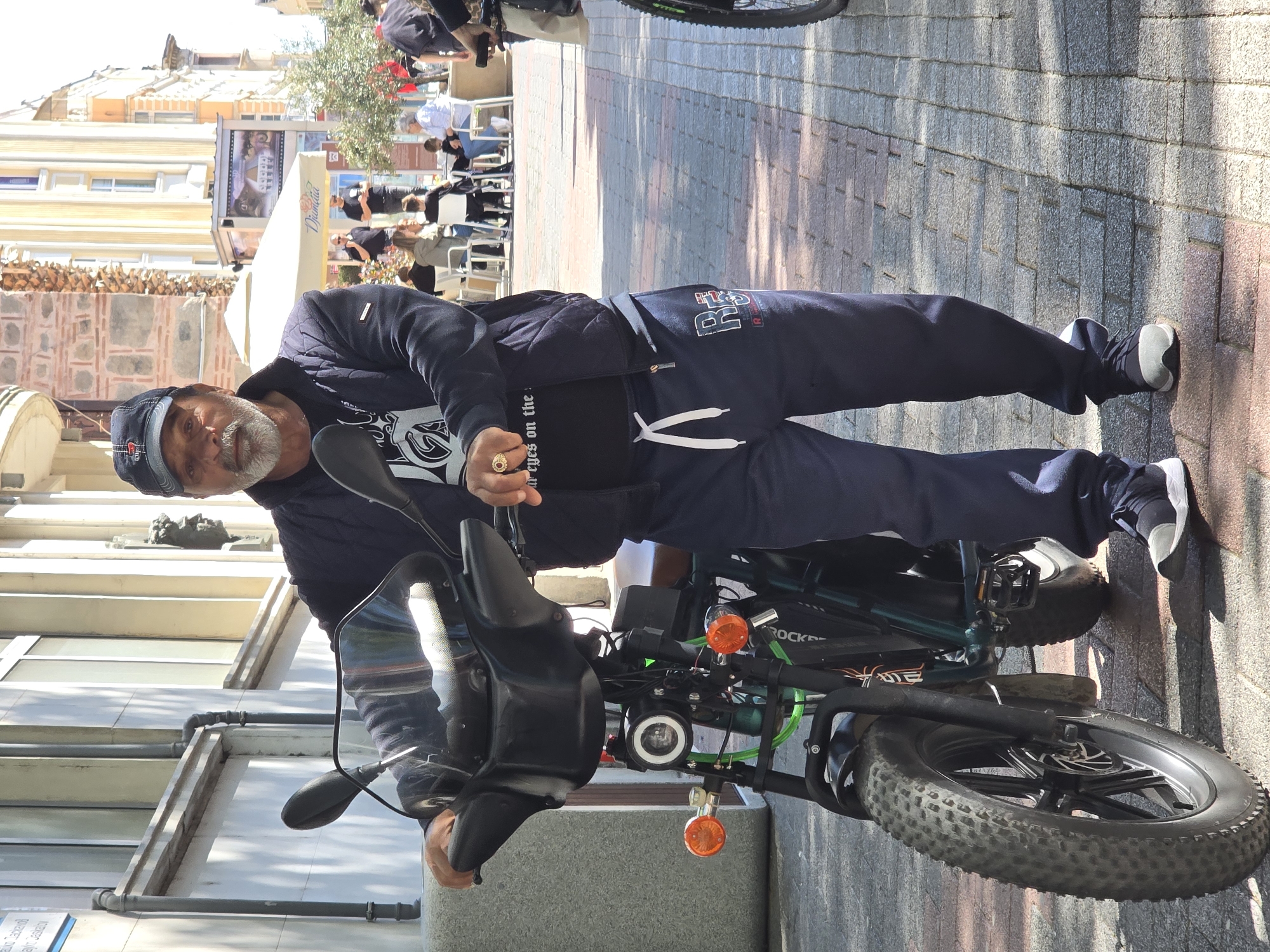A person is positioned next to a black bicycle. The person wears a dark blue cap, a dark jacket, and dark blue pants. They are on a paved surface that appears to be a street or sidewalk. In the background, there are buildings, some of which are multi-story, and people seated at tables, suggesting an outdoor café or restaurant area. The location appears to be in an urban environment, city of Unknown, Unknown.